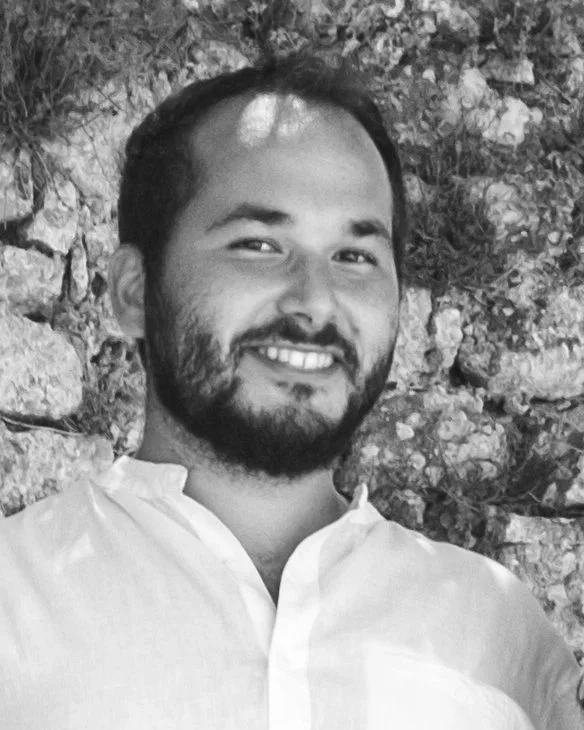 Black and white photo of a smiling man with a beard, wearing a white shirt, standing outdoors in front of a stone wall.
