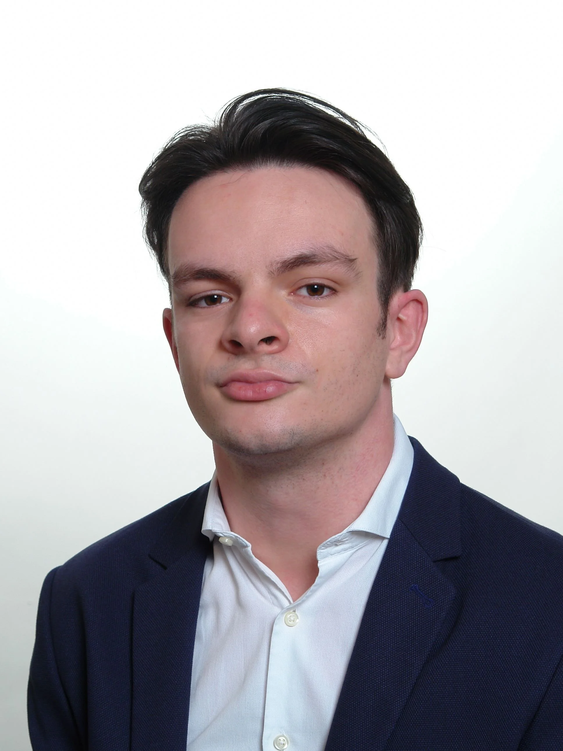Close-up of a young man wearing a dark blazer and white dress shirt, with a neutral expression, against a plain white background.