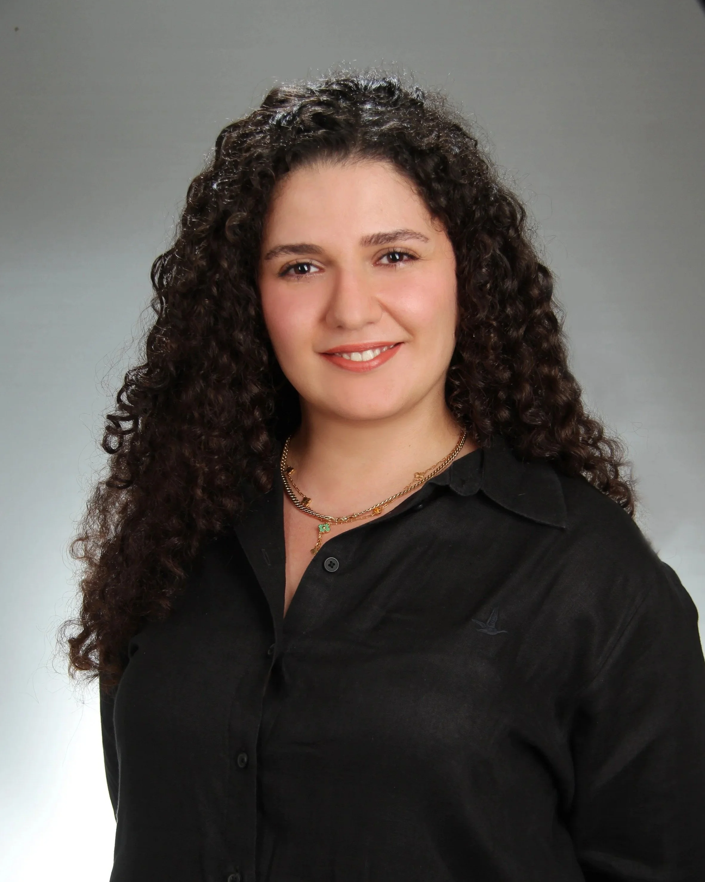 A woman with long curly dark hair wearing a black shirt and gold jewelry, smiling against a plain gray background.