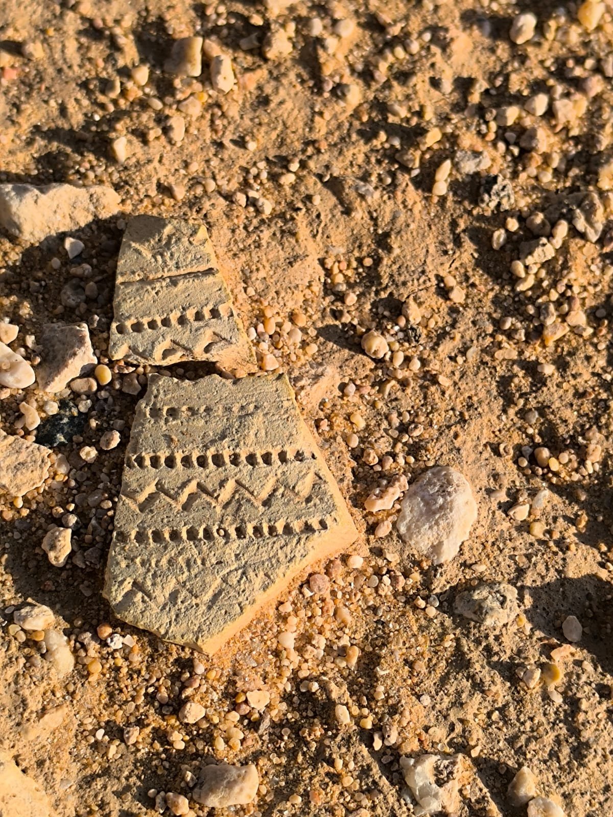 Close-up of sandy ground with small pebbles and broken piece of pottery with decorative patterns.