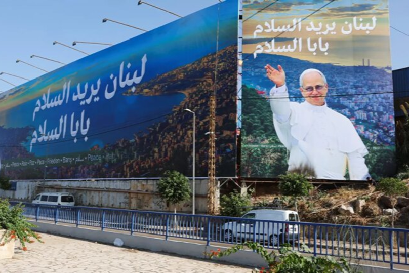 Large billboard in Lebanon featuring a portrait of a man waving, with text in Arabic that reads "Lebanon wishes peace" over a mountainous landscape background.