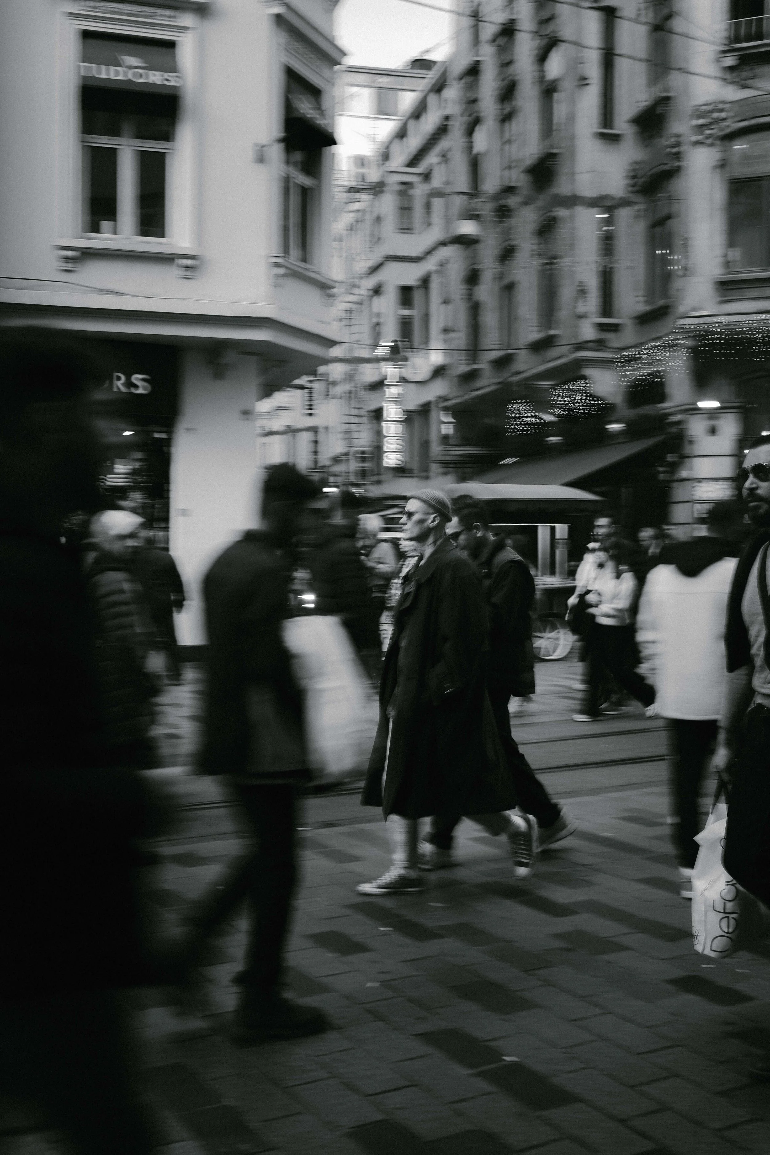 Black and white photo of people walking on a city street with buildings and shops in the background.