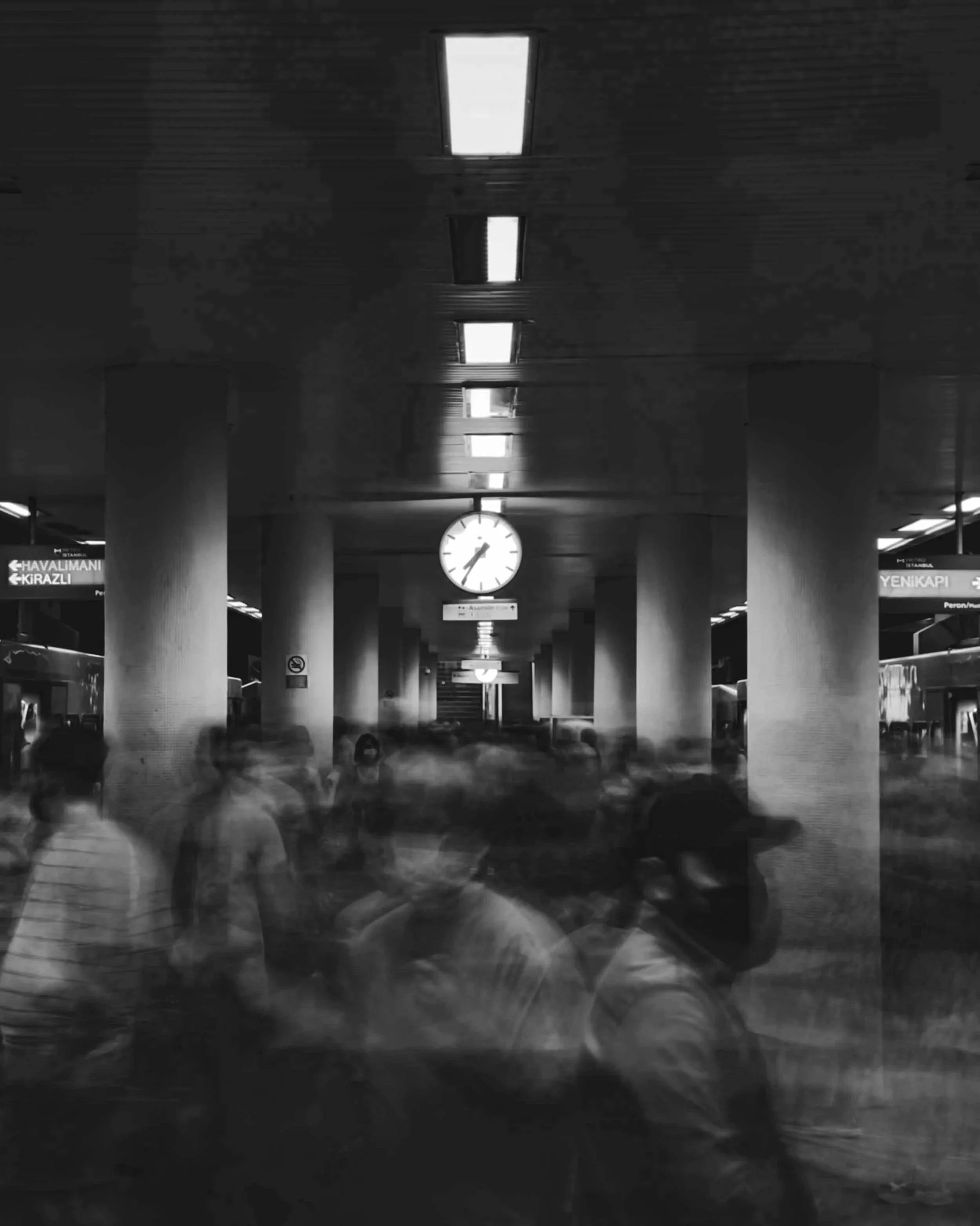 Black and white photo of an indoor space with a ceiling light, a clock, and blurry figures walking in the foreground.