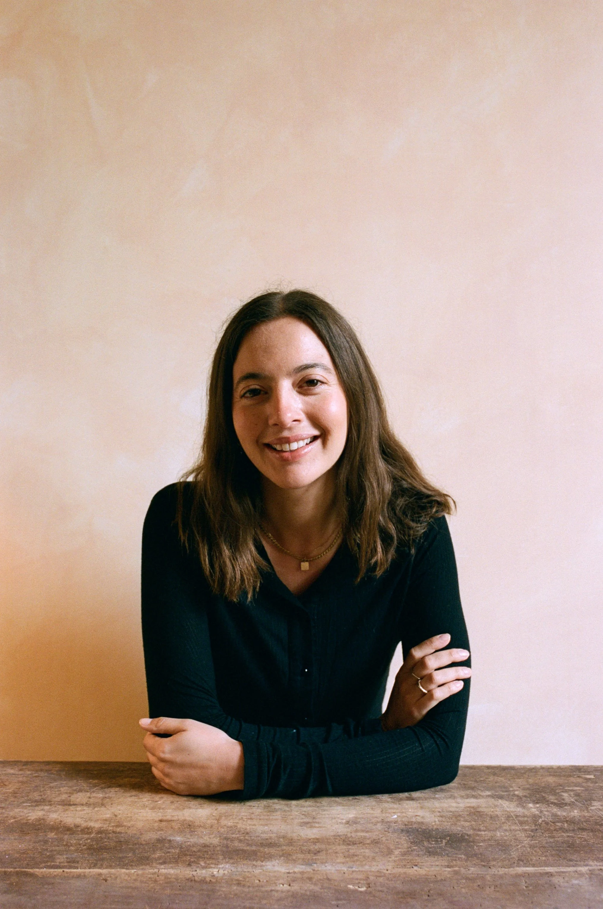 The therapist with long brown hair, wearing a black top and gold jewelry, sitting at a wooden table against a beige wall, smiling at the camera.