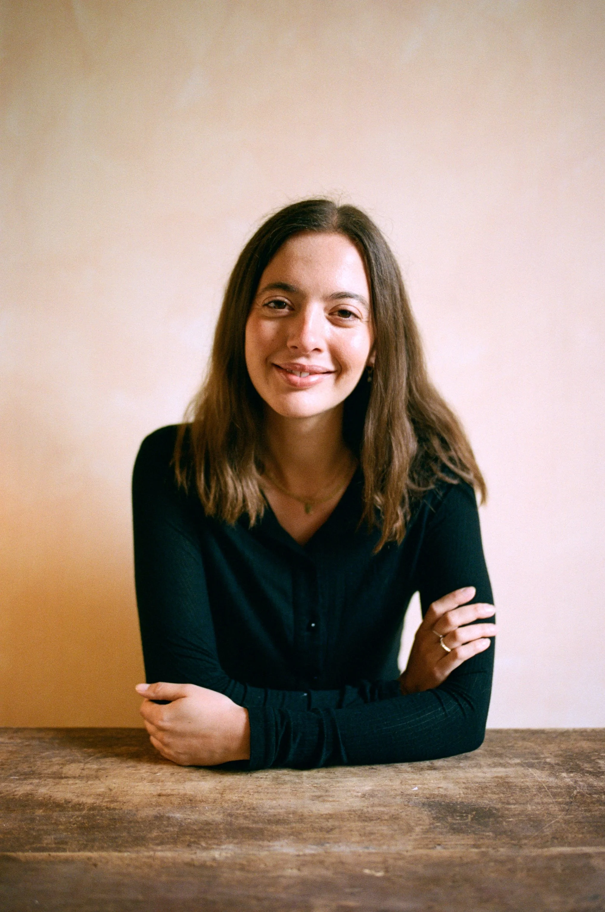 Portrait of a the therapist with long brown hair, wearing a black long-sleeve top, seated at a rustic wooden table against a beige wall.