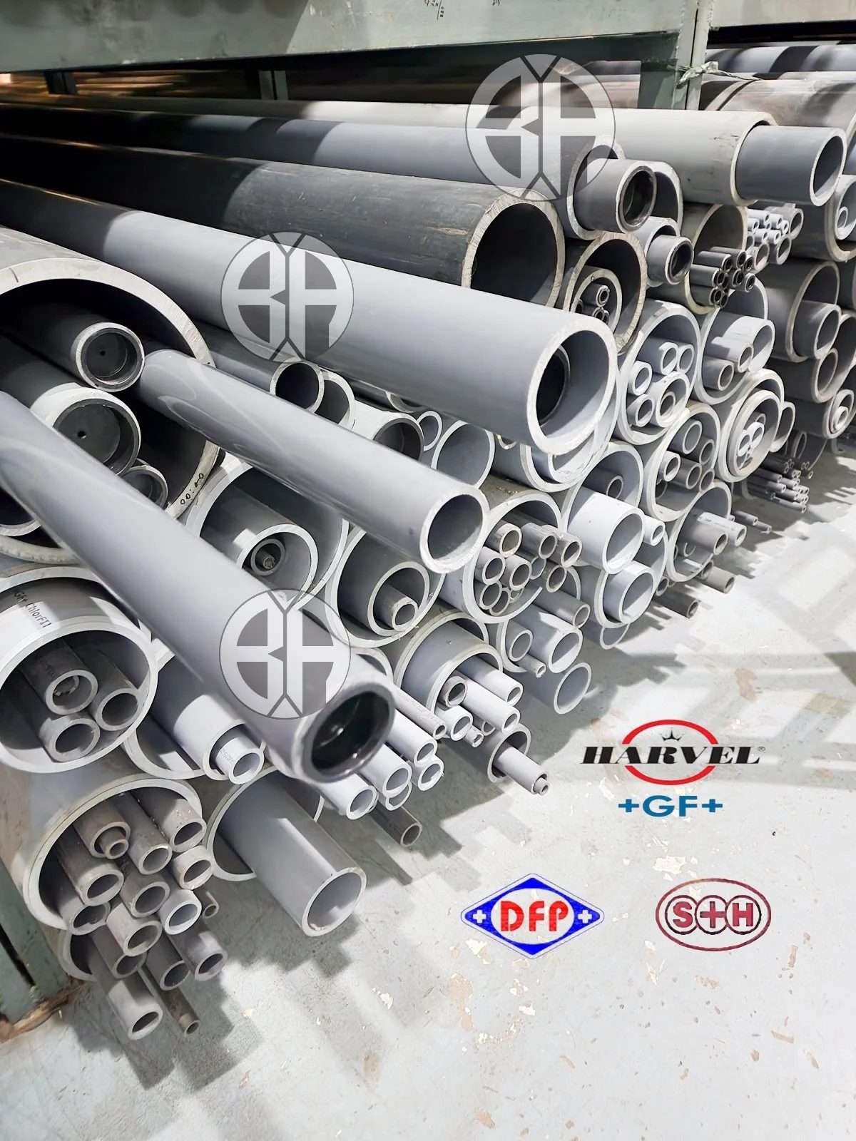 A stack of various metal pipes and tubes of different sizes stored outdoors on a metal rack. There are logos of different brands or companies, such as HARVEL, +GF+, DFP, and S+H, visible on the ground in front of the pipes.