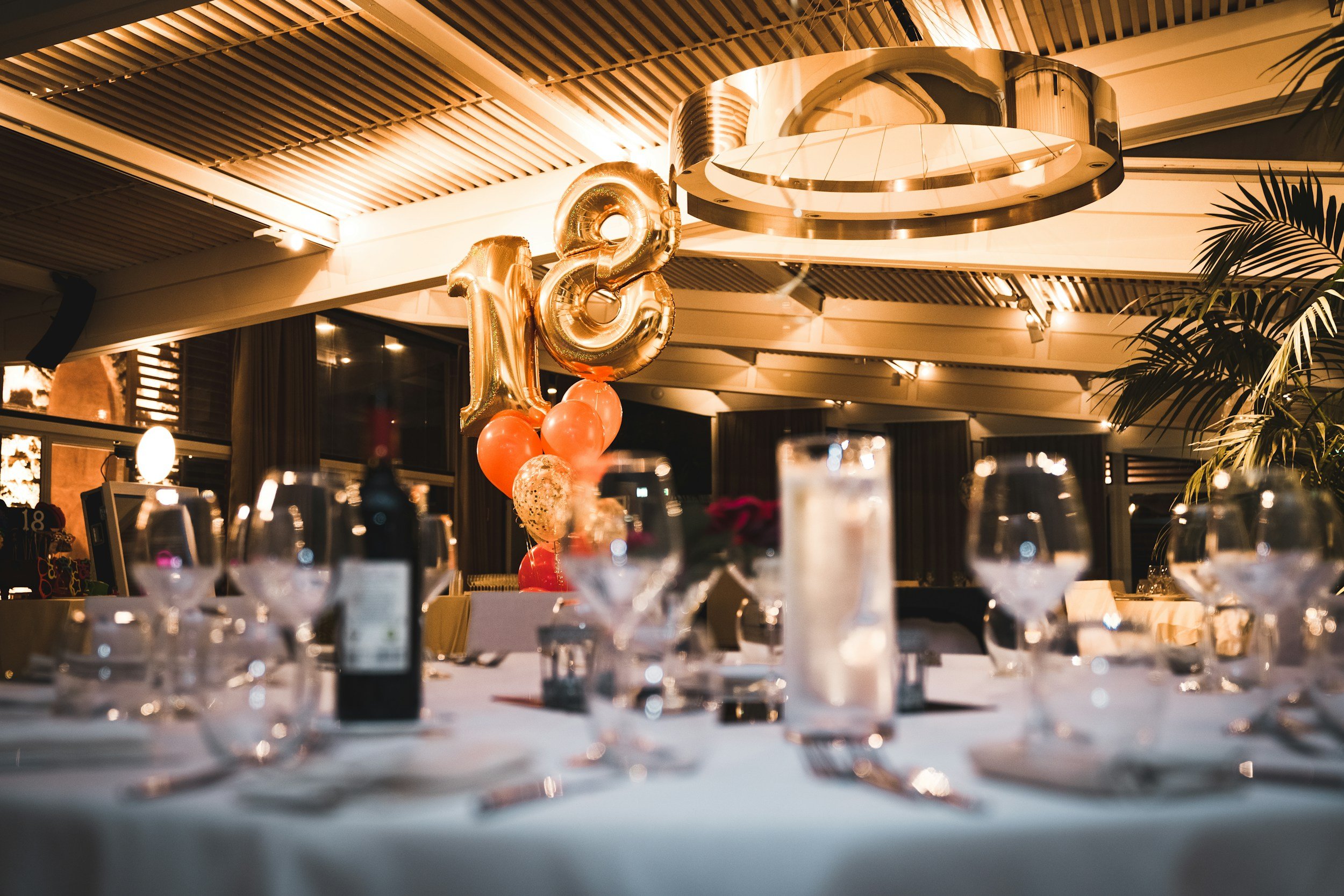 Une salle de fête décorée pour une célébration d'anniversaire avec des ballons dorés formant le chiffre 12, une décoration élégante, et des tables prêtes pour un dîner, dans un intérieur moderne avec un plafond en lamelles en bois et un luminaire métallique.