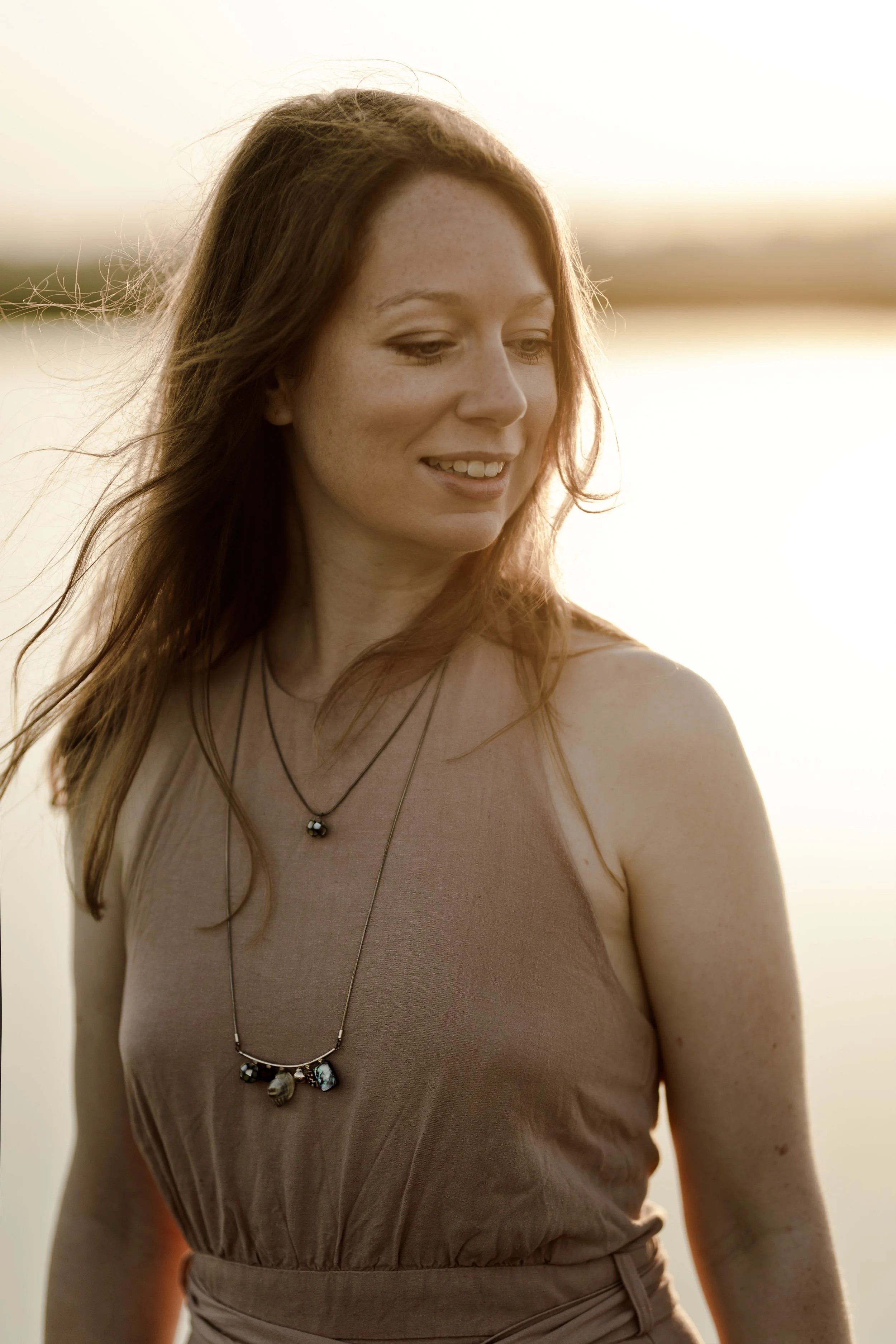 Jeune femme aux cheveux roux et peau claire, portant un haut sans manches de couleur beige, avec des colliers en pendentifs, regardant vers la droite, en arrière-plan un paysage flou lors du coucher du soleil.