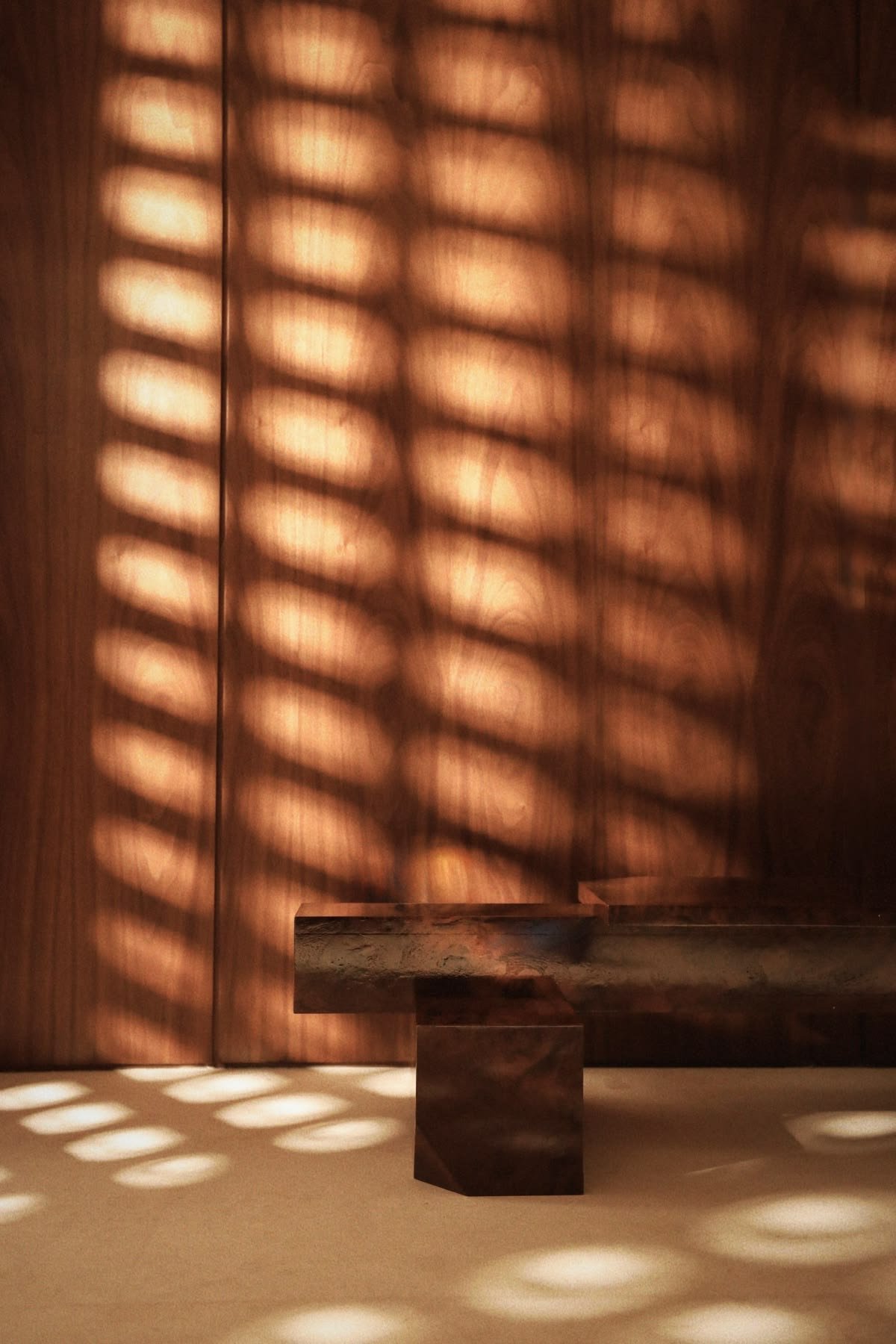 Ombres de stores ou fenêtres projetant des motifs en cercle sur un mur en bois et un sol, créant un jeu de lumière et d'ombres dans une pièce intérieure avec un meuble en pierre ou marbre.
