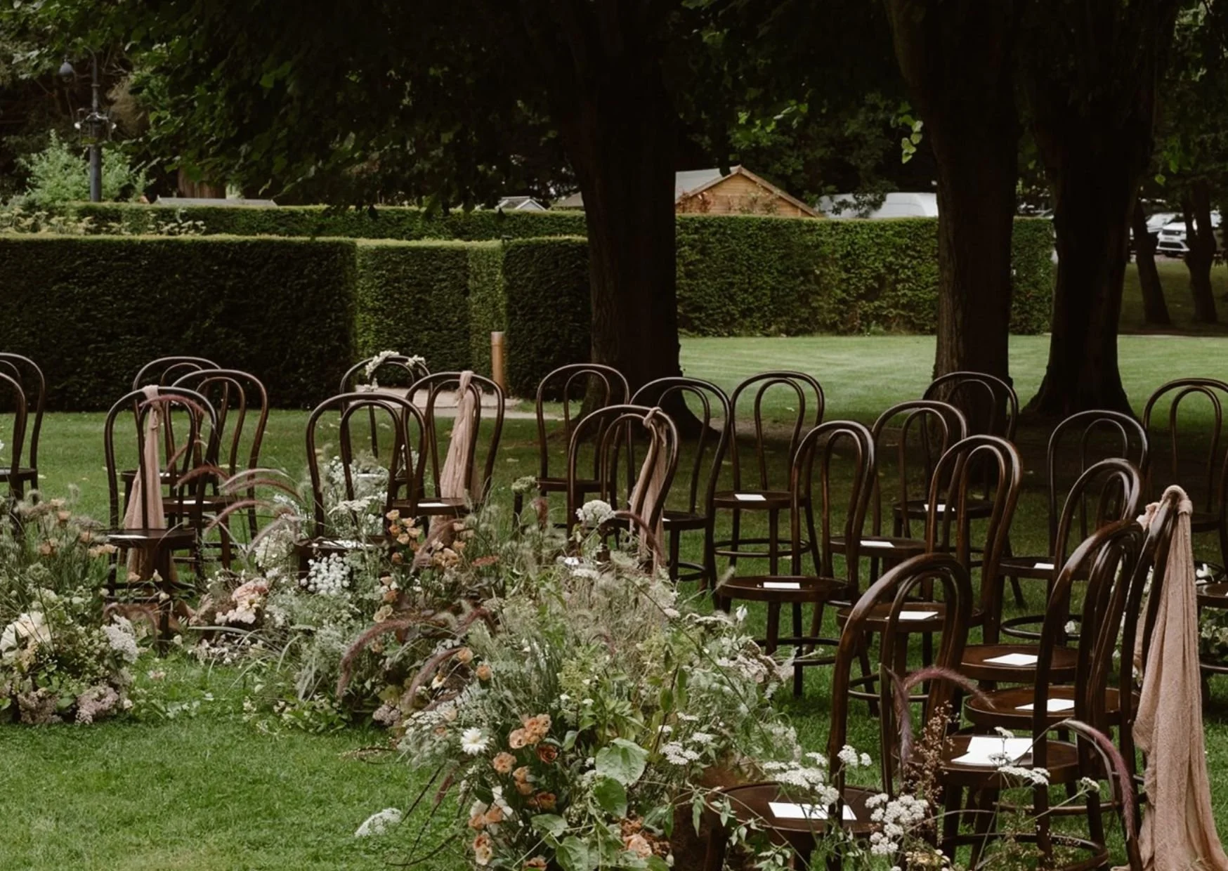 Outdoor wedding setup with wooden chairs and floral decorations on a grassy lawn, surrounded by trees and hedges.