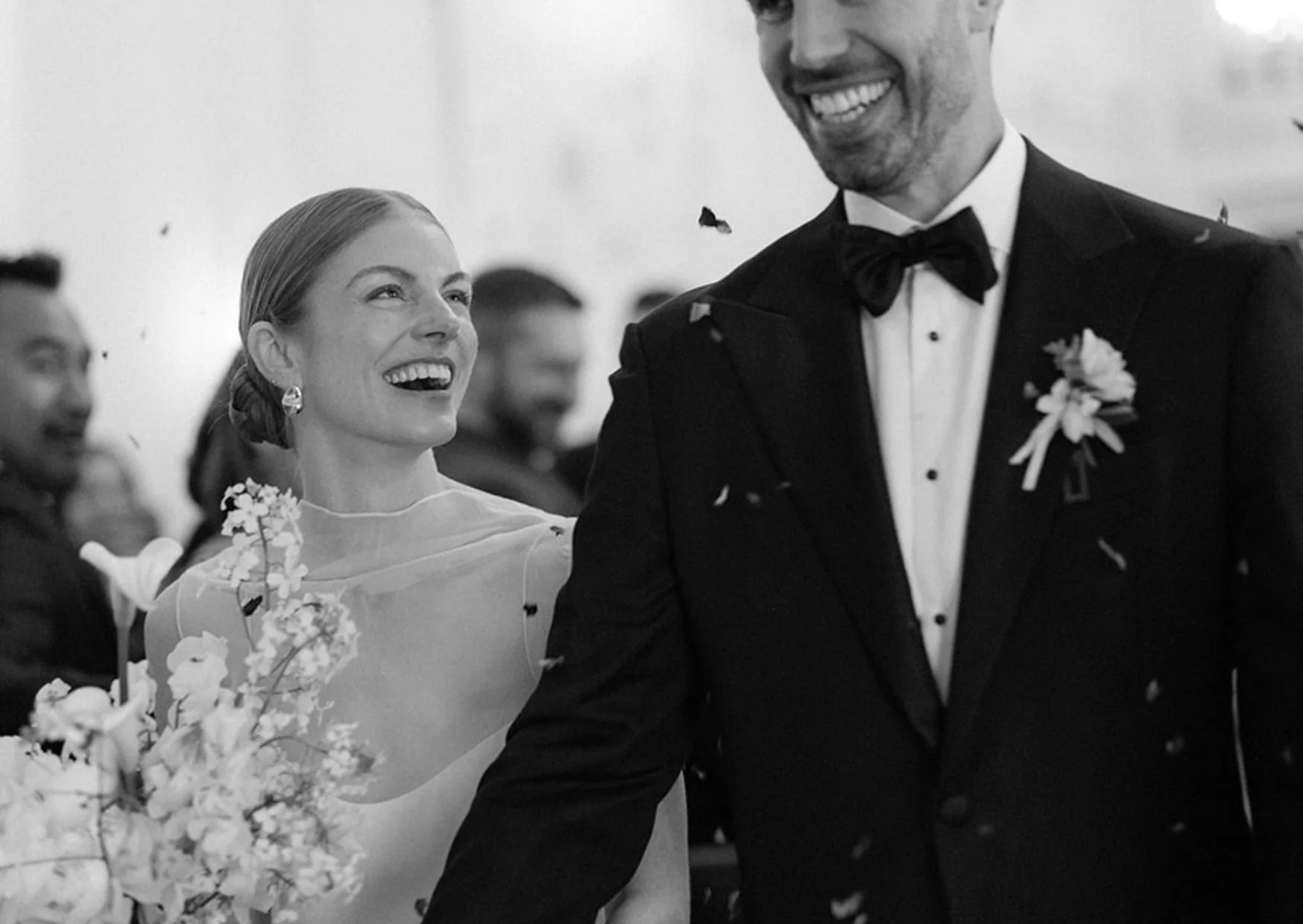 Black and white photo of a wedding celebration. A smiling woman in a wedding dress holding a bouquet looks at a smiling man in a tuxedo. Guests can be seen in the background with confetti falling.