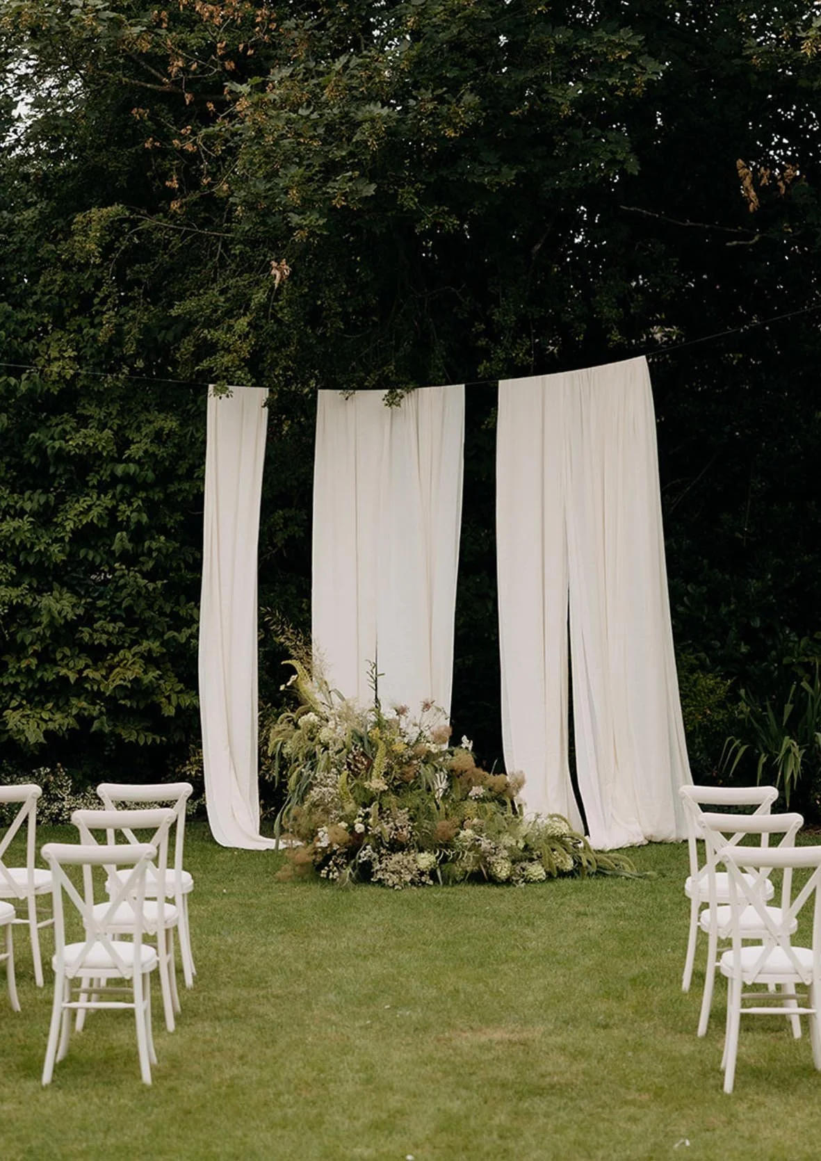 Outdoor wedding altar with white drapes, a floral arrangement at the base, and white chairs set up on a grassy lawn surrounded by trees.