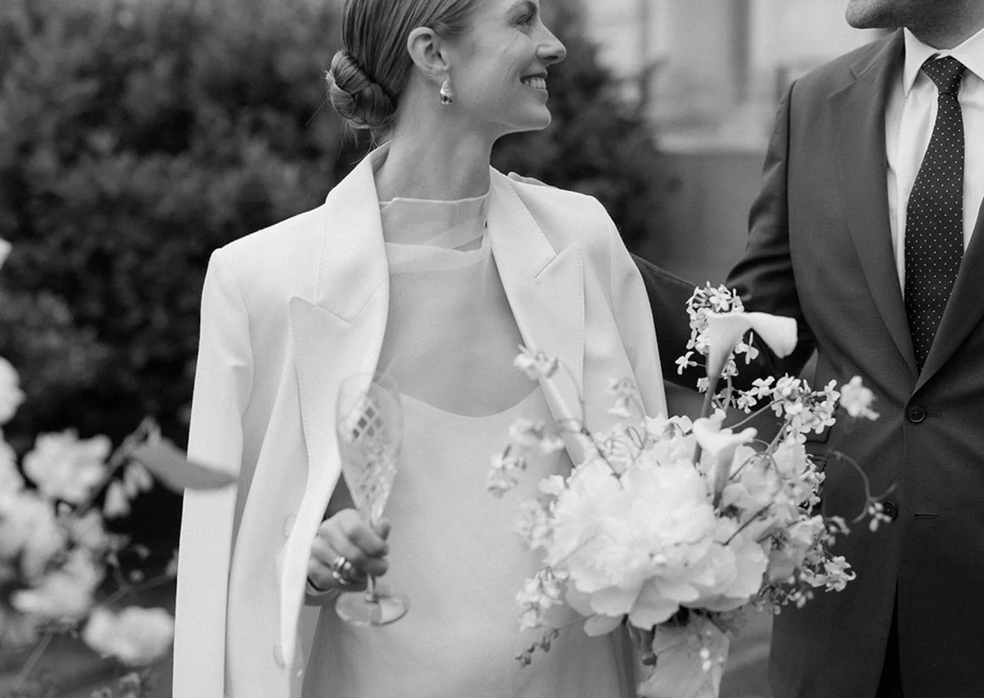 Black-and-white photo of a woman in a wedding dress holding a bouquet, smiling and looking at a man in a suit, outdoors with flowers and trees in background.