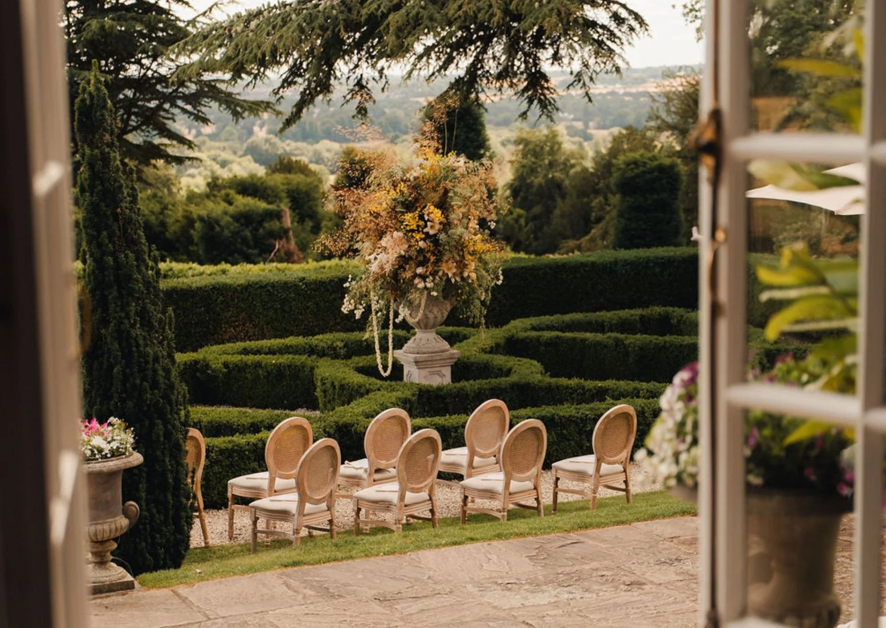 A garden scene viewed from an open window, featuring a long table with chairs, a large urn with a floral arrangement, neatly trimmed hedges, and lush trees in the background.