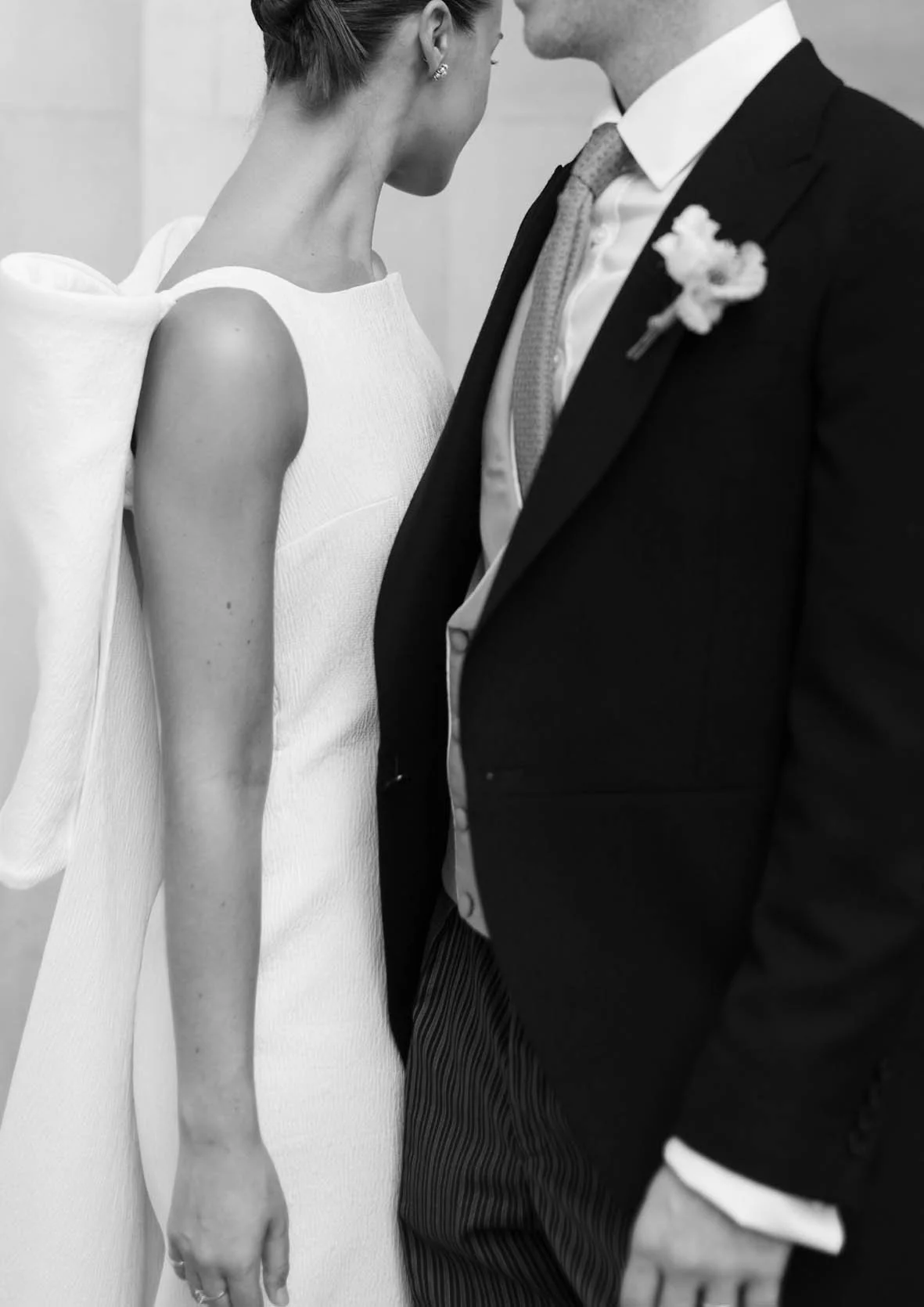 A black-and-white photo of a woman and a man close to each other, with their faces nearly touching. The woman wears a sleeveless white dress with a textured fabric, and the man is dressed in a dark suit with a white shirt, a patterned tie, and a floral boutonniere.