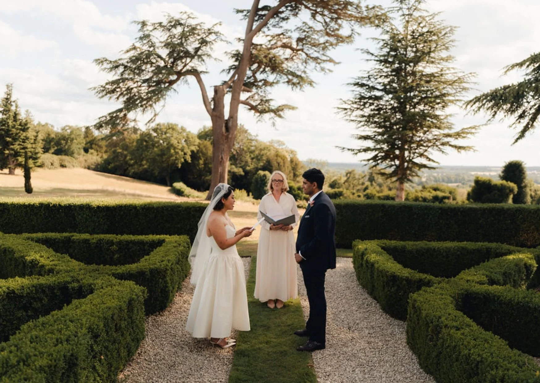A wedding ceremony outdoors in a garden with two people dressed in wedding attire standing in front of an officiant, exchanging vows while an officiant holds a book. The scene is set among neatly trimmed green hedges with trees and rolling landscape in the background.
