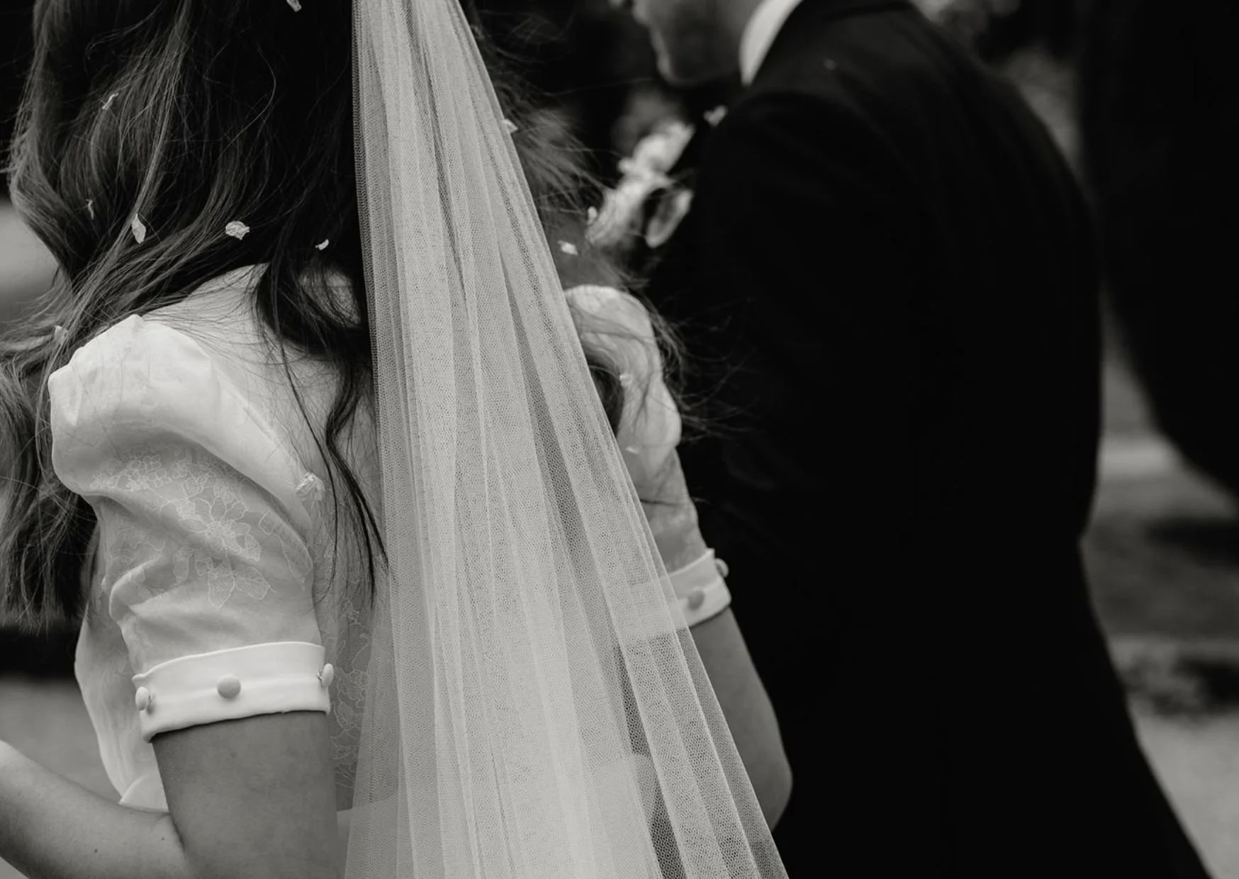 Black and white photo of a bride and groom holding hands during a wedding ceremony, with part of the bride's veil and dress visible.