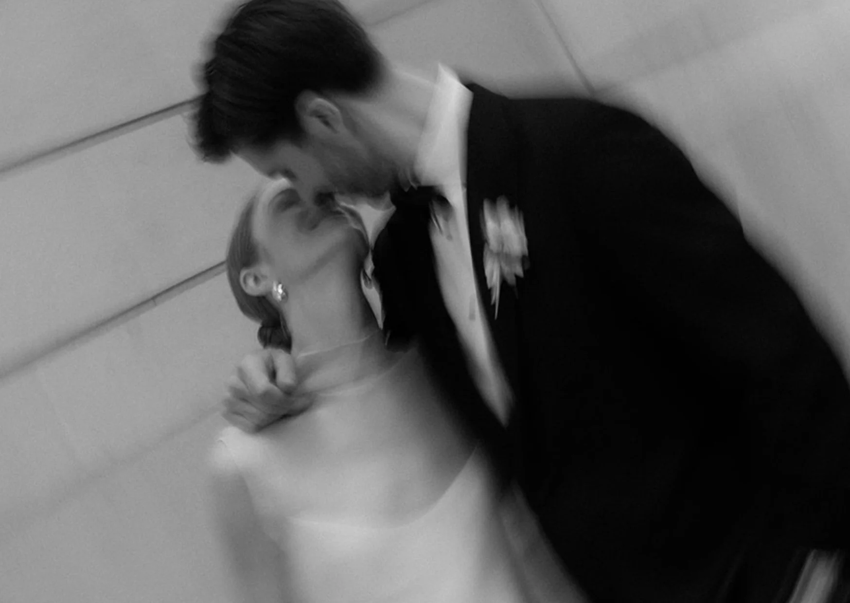 A man in a tuxedo and a woman in a wedding dress share a kiss, with the woman holding the man's face and the man holding her waist, against a plain wall.