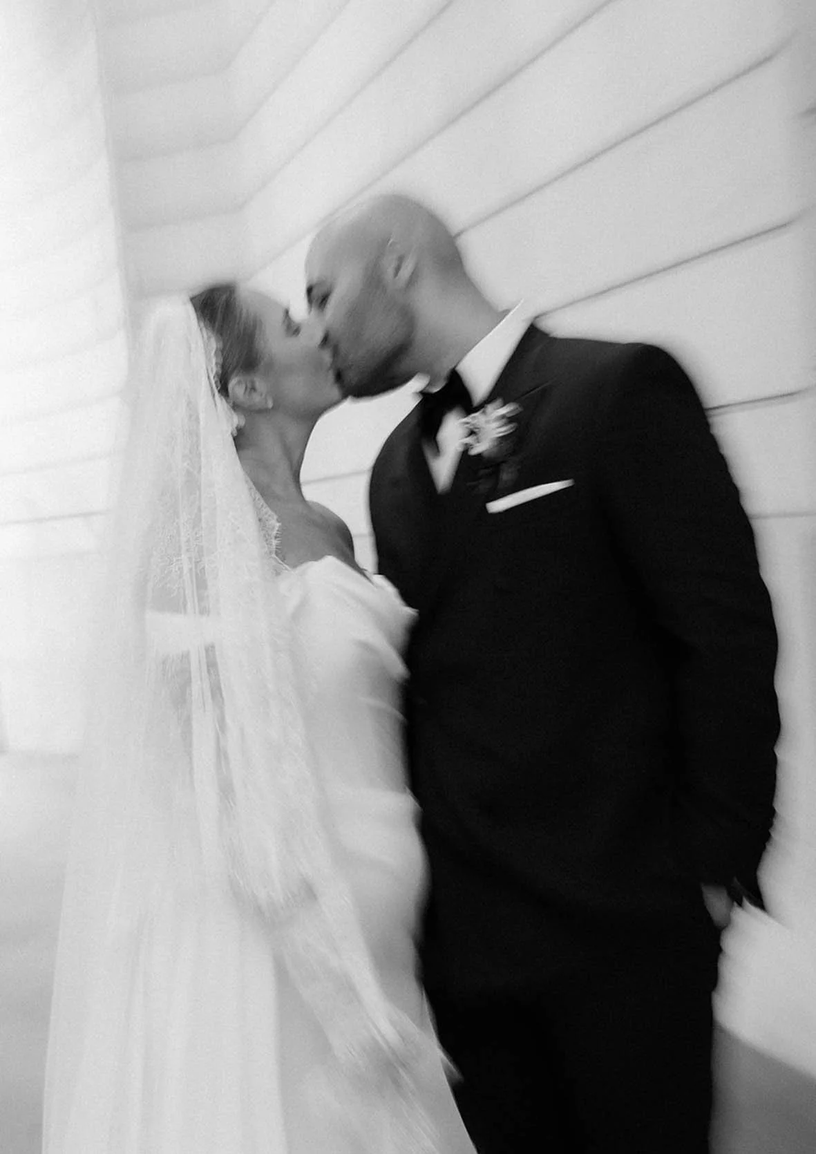 Black and white photo of a bride and groom kissing indoors, against a wooden wall. The bride is wearing a white wedding dress and veil; the groom is in a black tuxedo with a bow tie.