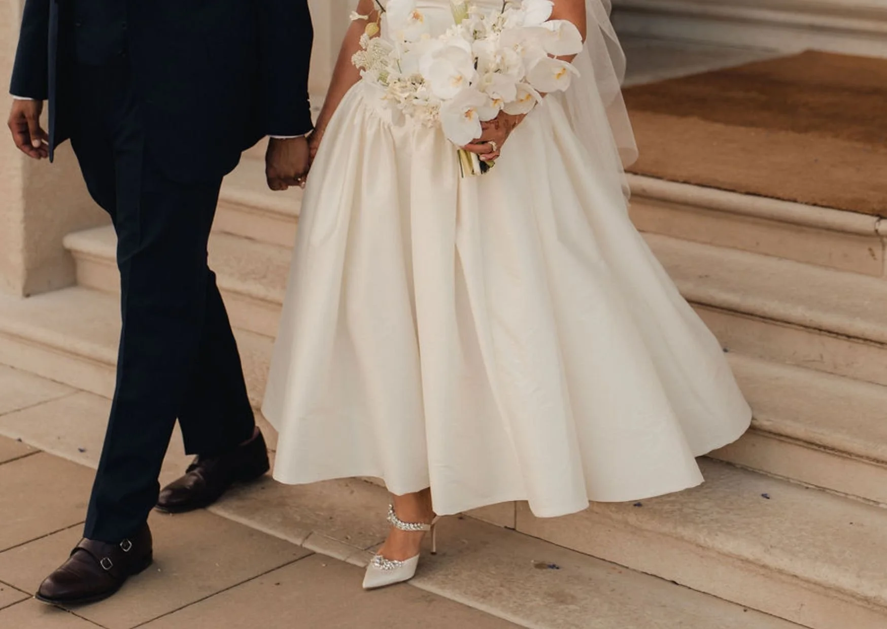 A bride in a white gown holding a bouquet of white flowers and a groom in a dark suit standing together on the steps of a building.