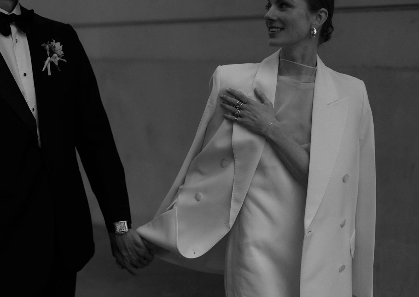 Woman in a white suit smiling and looking to the side, holding hands with a person in a tuxedo.
