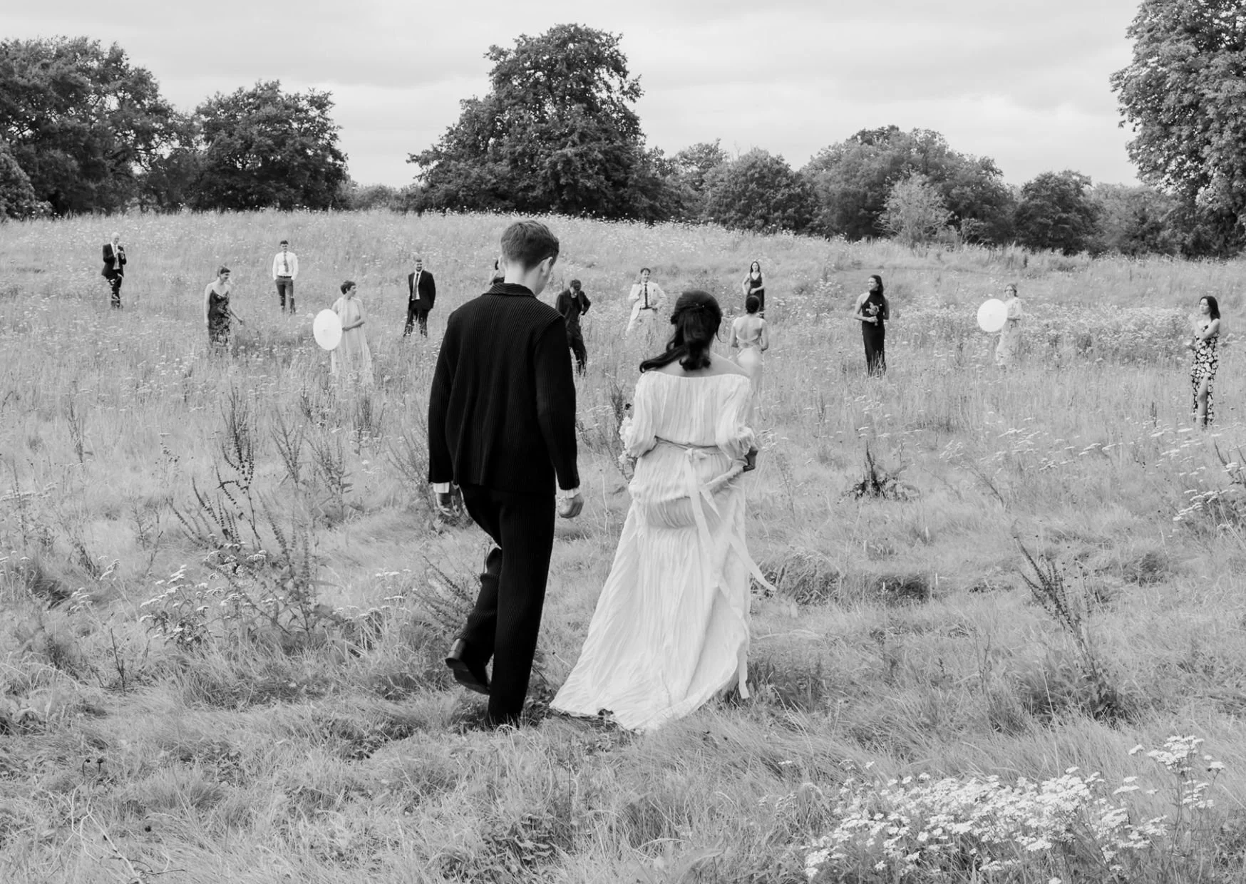 A black and white photo of a wedding ceremony outdoors in a grassy field with trees in the background. The bride and groom walk away from the camera, with the groom in a dark suit and the bride in a long, flowing wedding dress. In the distance, there are guests standing and observing the ceremony, some holding balloons.