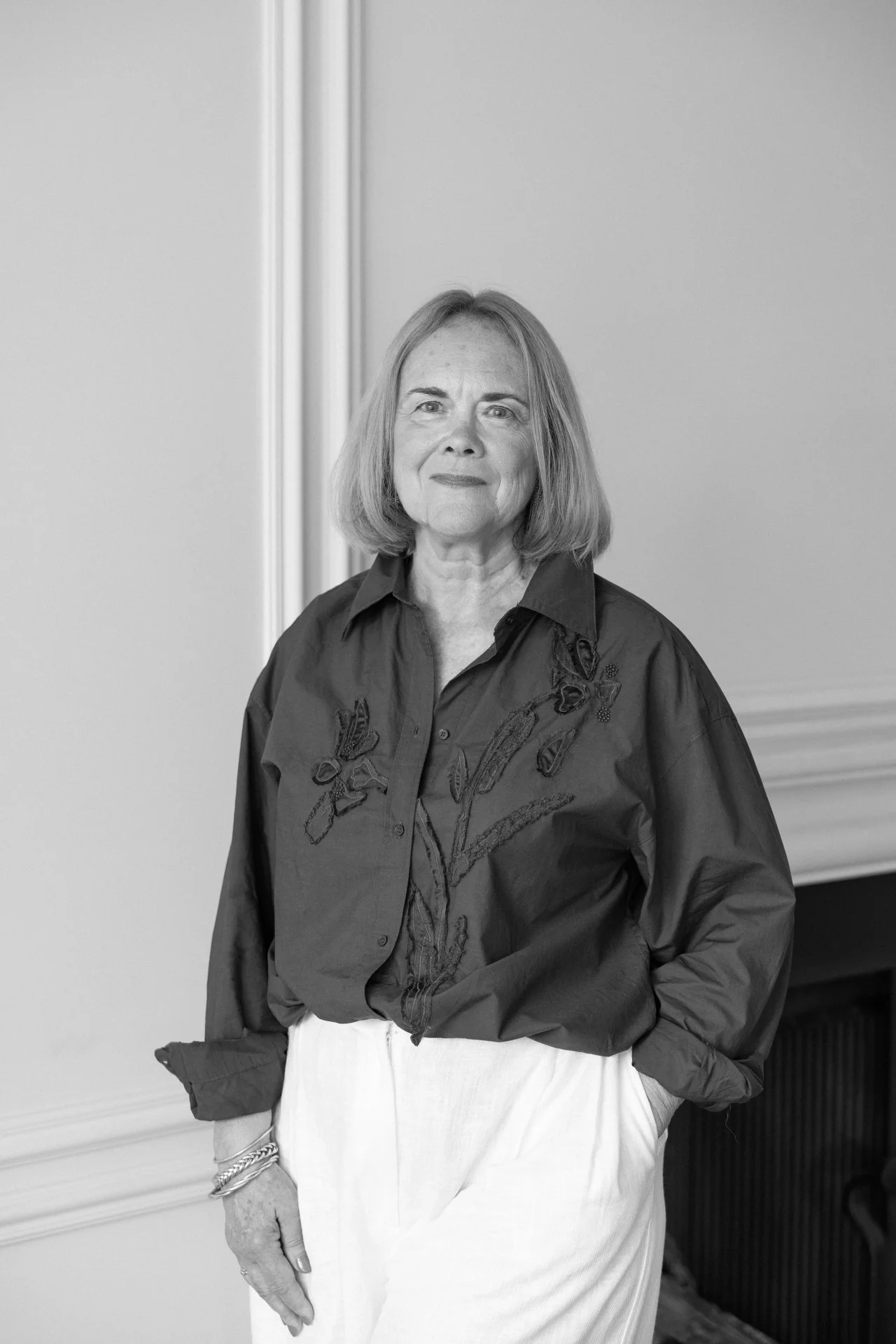 Black and white photo of an older woman with shoulder-length hair wearing a dark embroidered blouse and light-colored pants, standing indoors against a plain background.