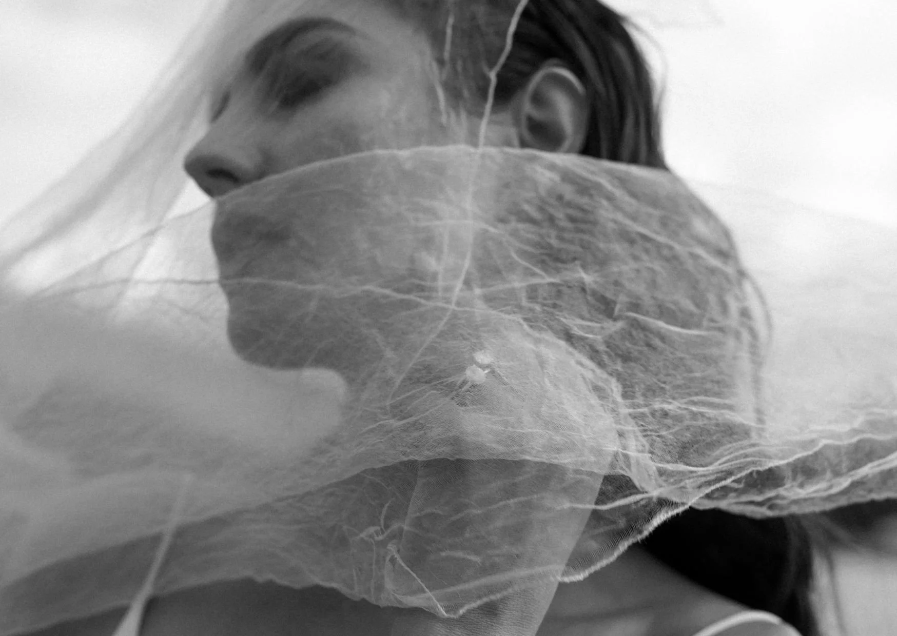 A person with dark hair wears a face mask and appears to be resting or sleeping, with a translucent veil or cloth in front of them.