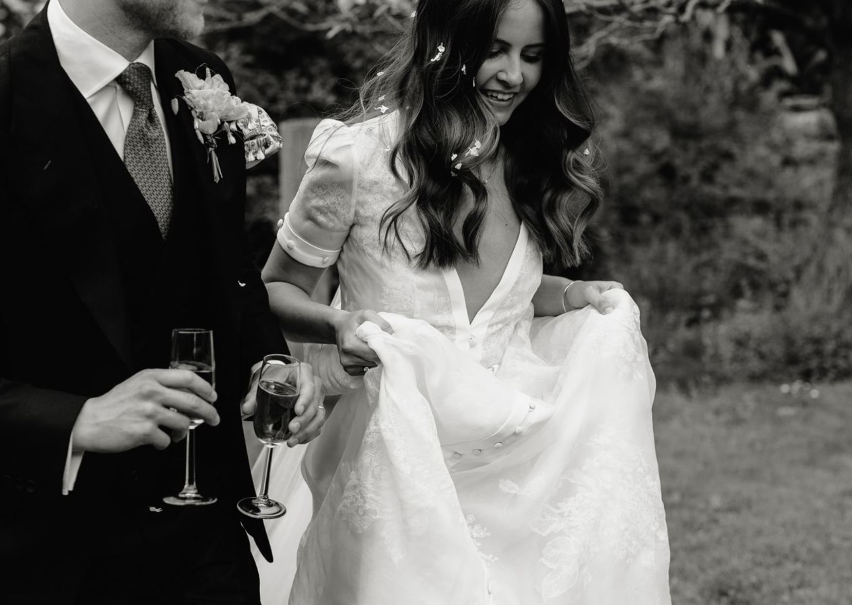 A black and white photo of a bride and groom holding champagne glasses at their wedding. The bride is smiling and wearing a wedding dress, while the groom is in a suit and tie. They are outdoors in a natural setting.