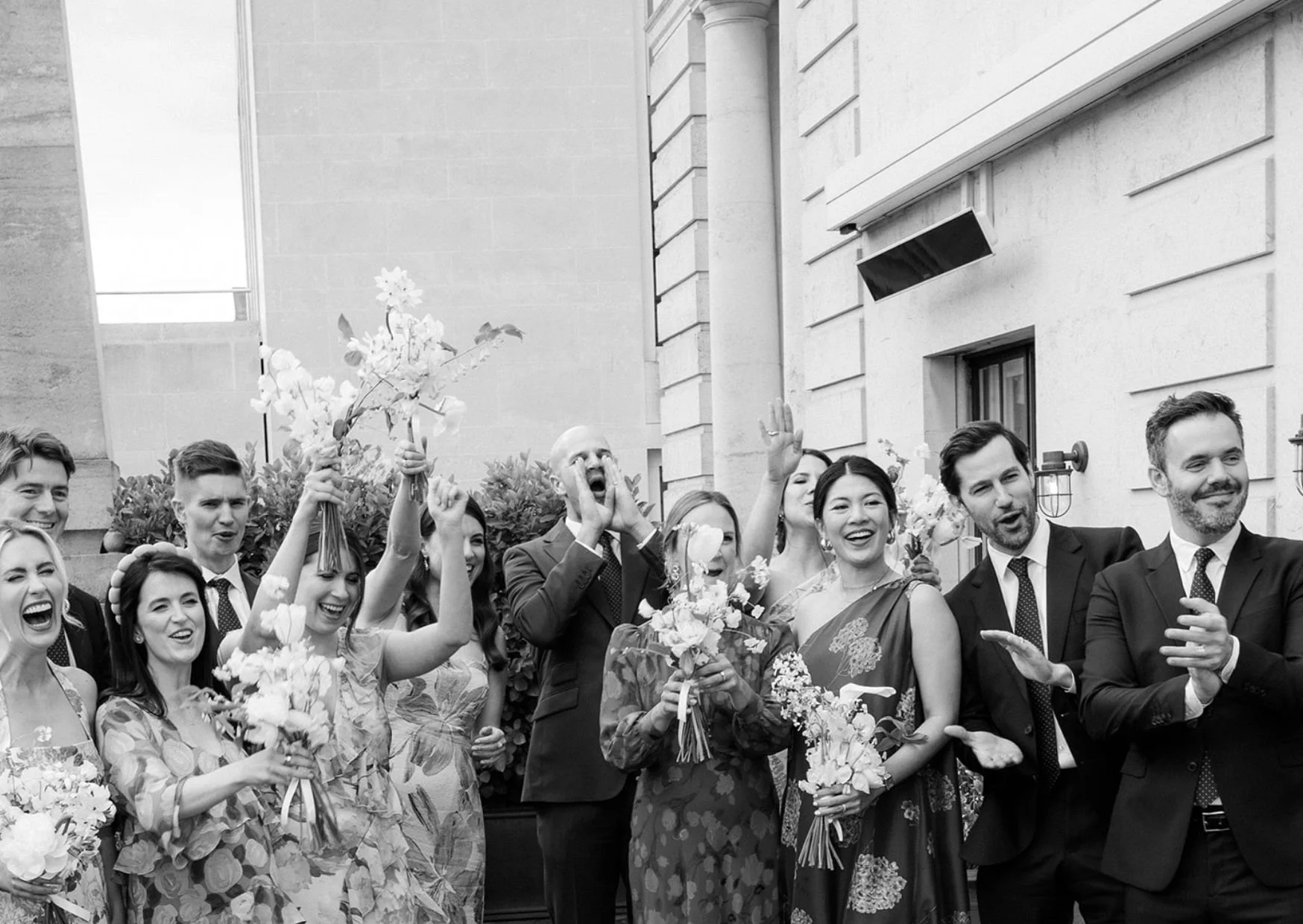 A group of people dressed in formal attire celebrating outdoors, holding bouquets, some smiling and gesturing excitedly, with a building and greenery in the background.