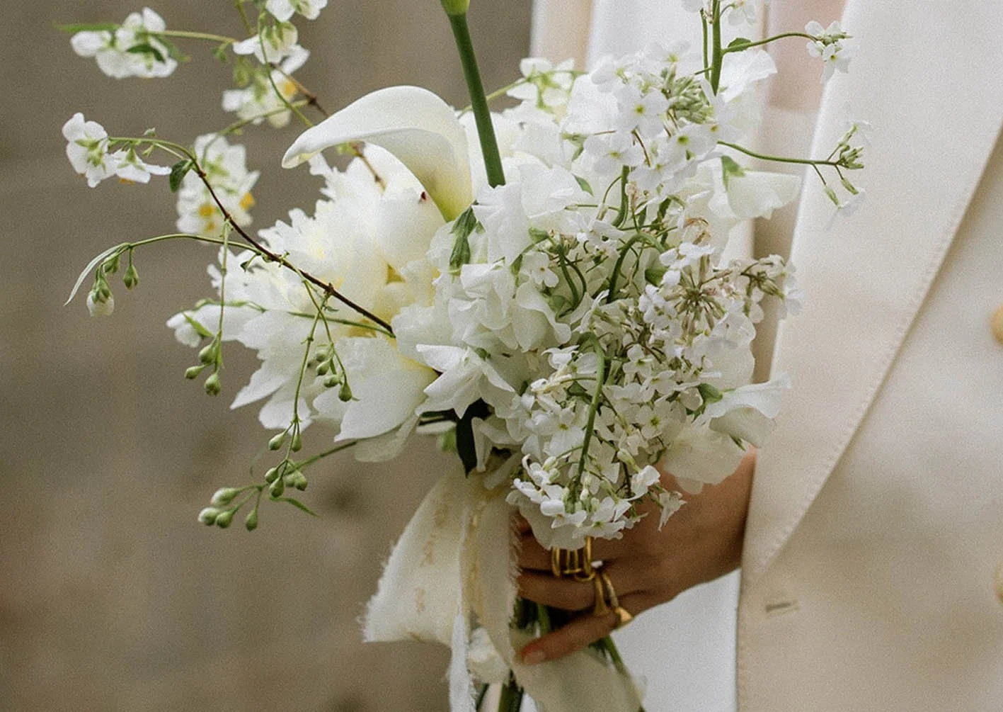 A hand holding a bouquet of white flowers, including lilies, roses, and smaller blooms, with some greenery, against a neutral background.