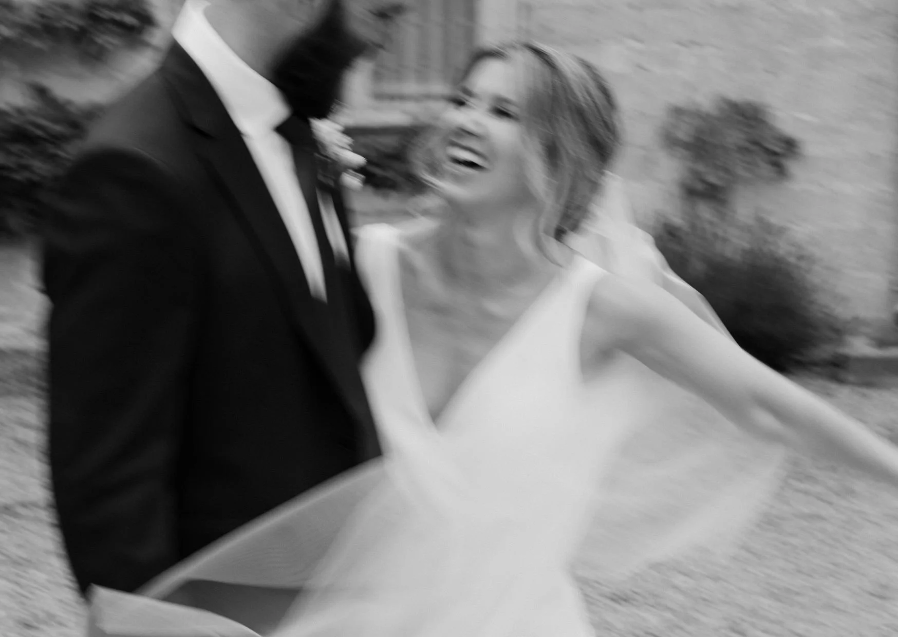 Black and white photo of a smiling woman in a white wedding dress and a man in a tuxedo, both looking at each other, outdoors with a brick wall background.