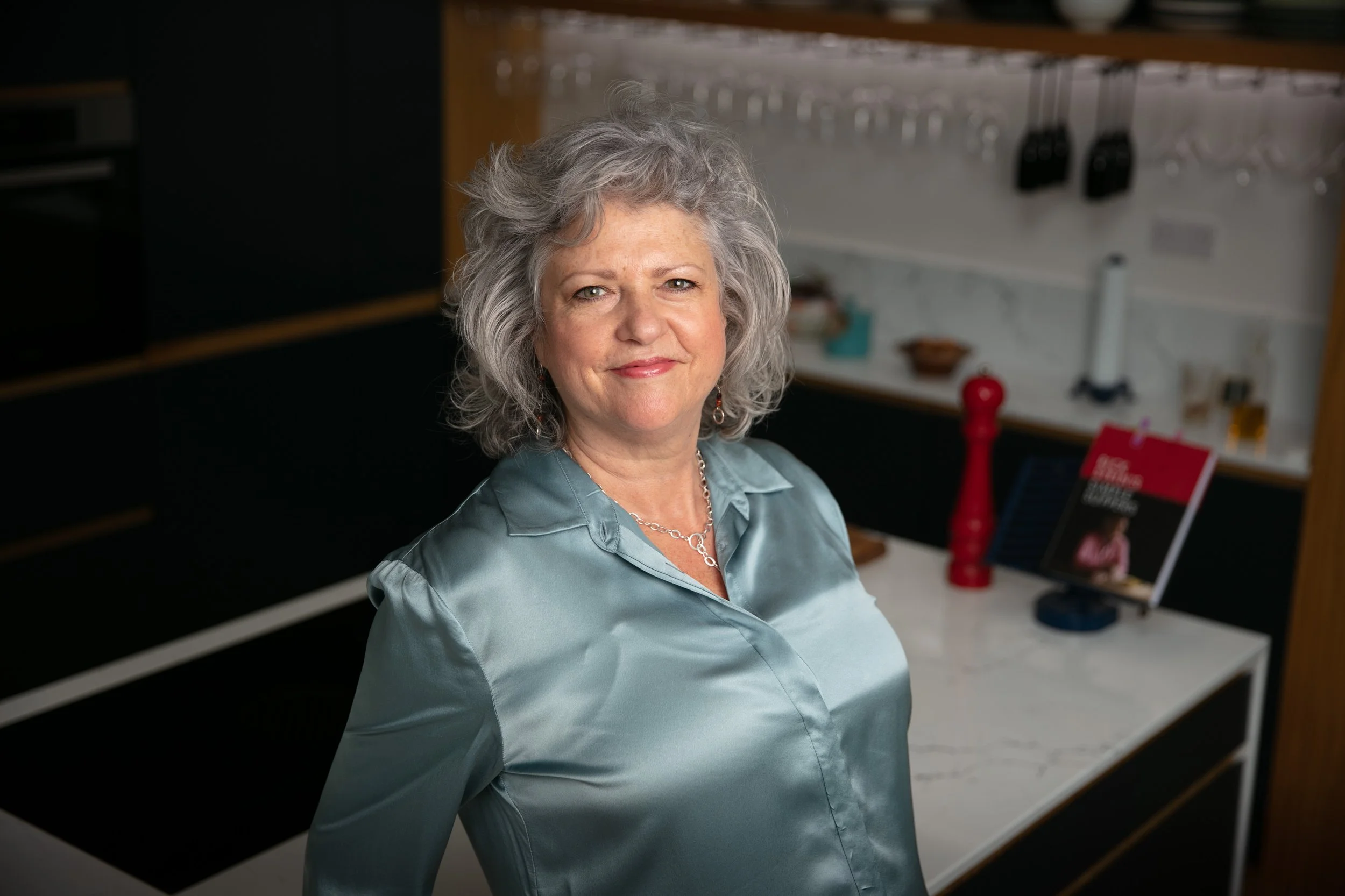 A smiling woman with short hair wearing a light blue satin blouse, standing in a kitchen with black and wooden cabinets and a countertop with kitchen utensils.