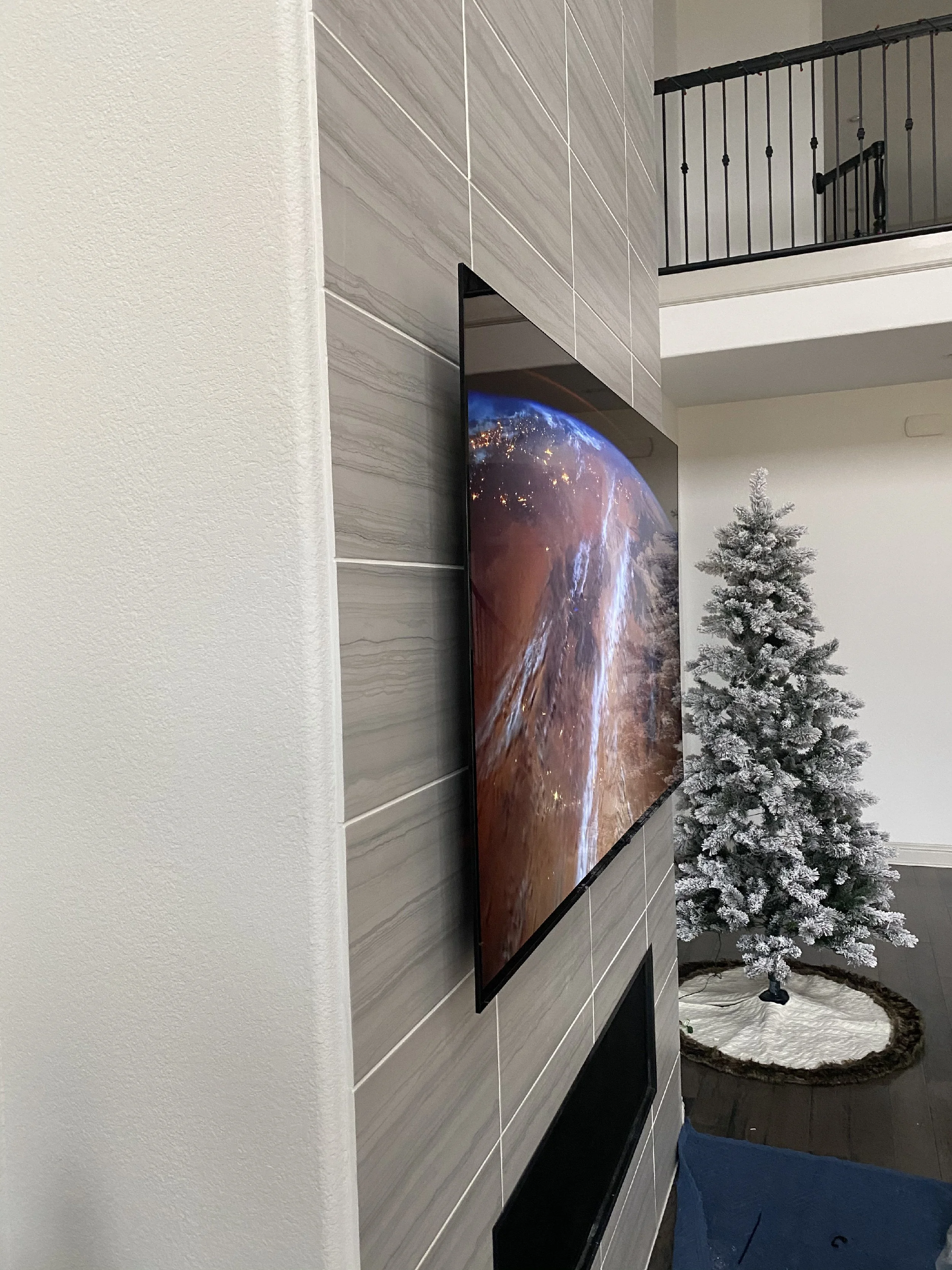 Indoor scene with a mounted flat-screen television on a wall, next to an artificial snow-covered Christmas tree, with a circular rug on the floor