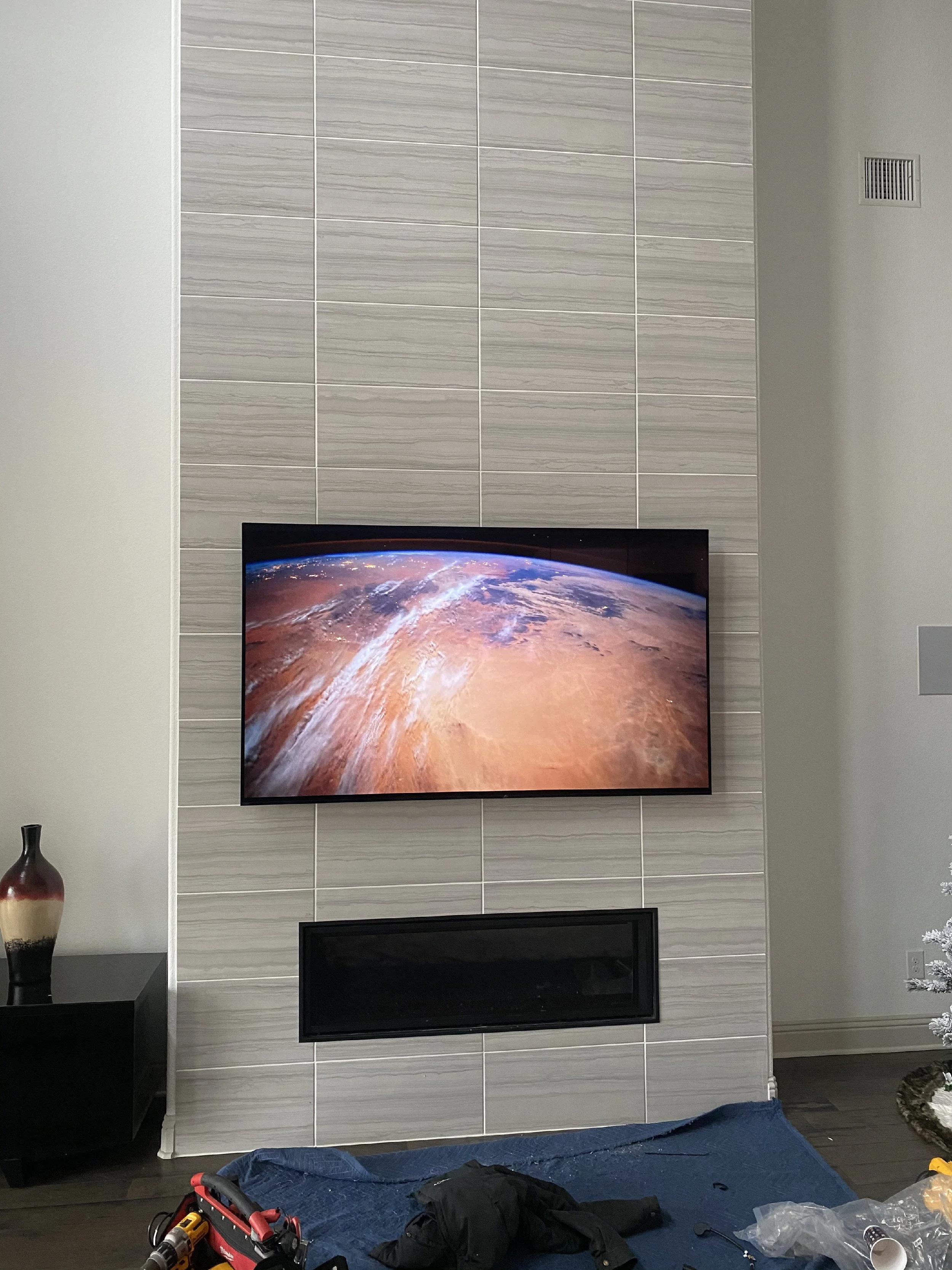 Living room with a mounted flat-screen TV showing Earth from space on a textured tile wall. Below the TV is an empty black fireplace, with a small black side table on the left holding a ceramic vase. In the foreground, there is a blue tarp with various tools and clothing laid on it, including a red toolbox, power drill, and plastic bags.