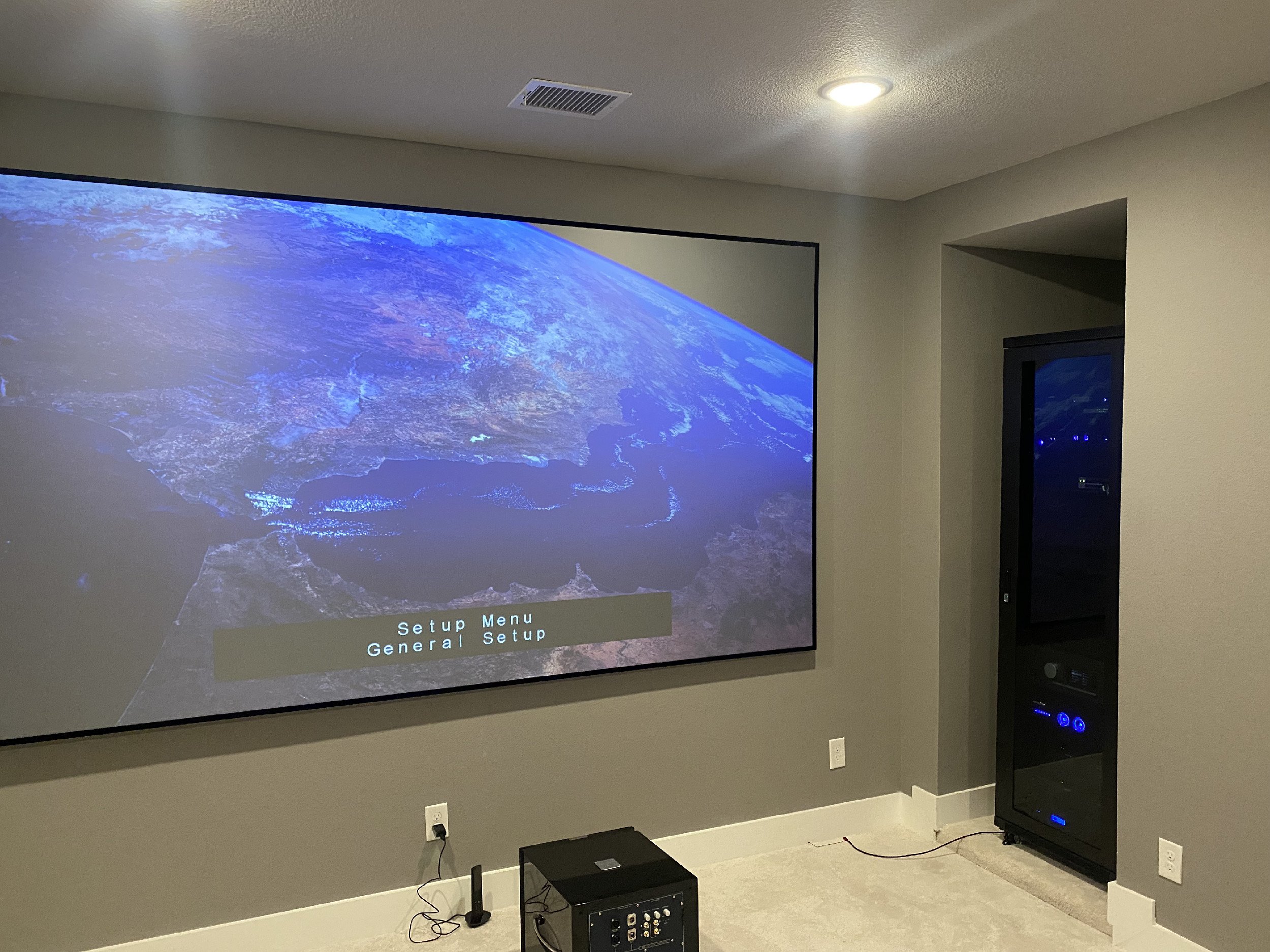 A large projector screen displaying an image of Earth from space in a media room with a gray wall, ceiling light, and a black entertainment rack.