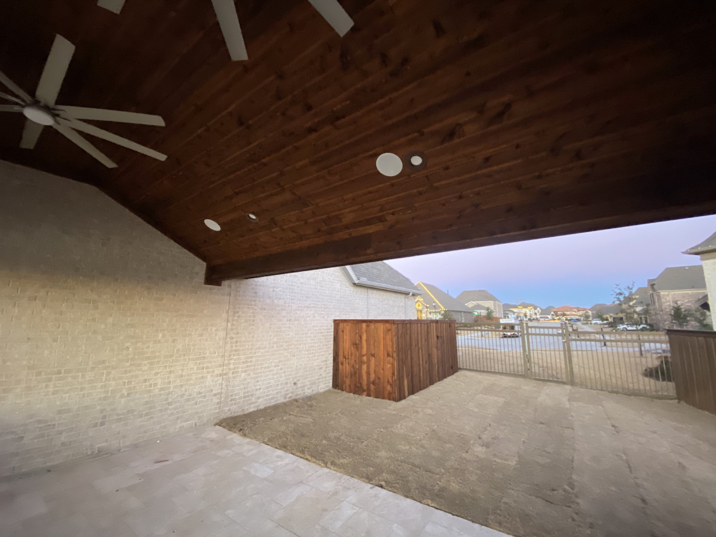 View of a backyard patio area with a brick wall, a wooden fence, and a covered wooden ceiling with recessed lights. The area is paved with stone tiles and is adjacent to neighboring houses with lit windows.