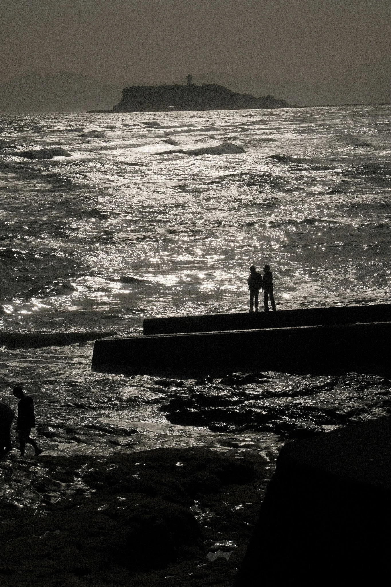 Silhouettes of people on a beach at sunset, ocean waves reflecting light, a distant island with a lighthouse on top, and mountains in the background.