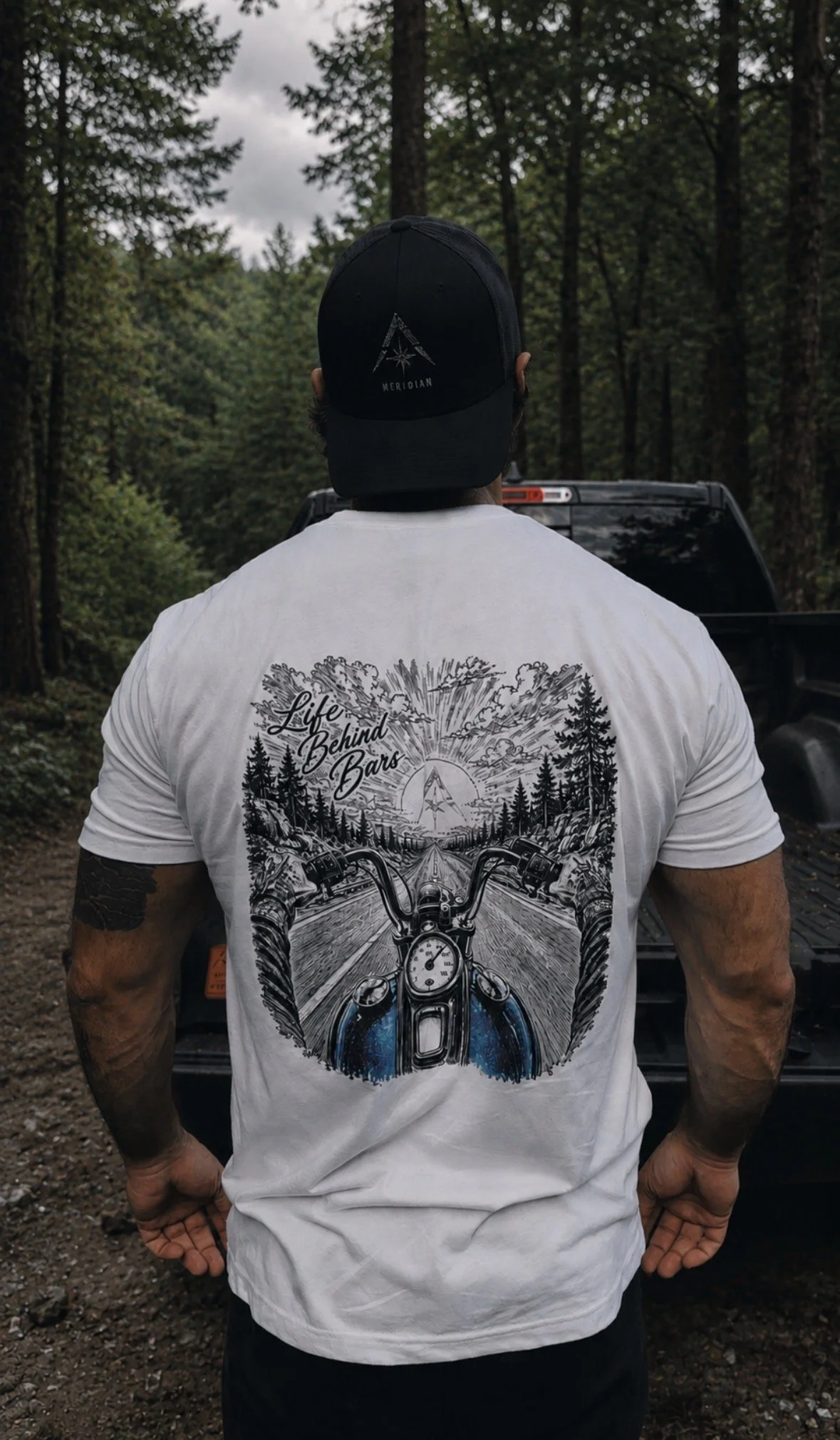 A man wearing a black cap and a white t-shirt with motorcycle art in a forested outdoor setting, facing away from the camera.
