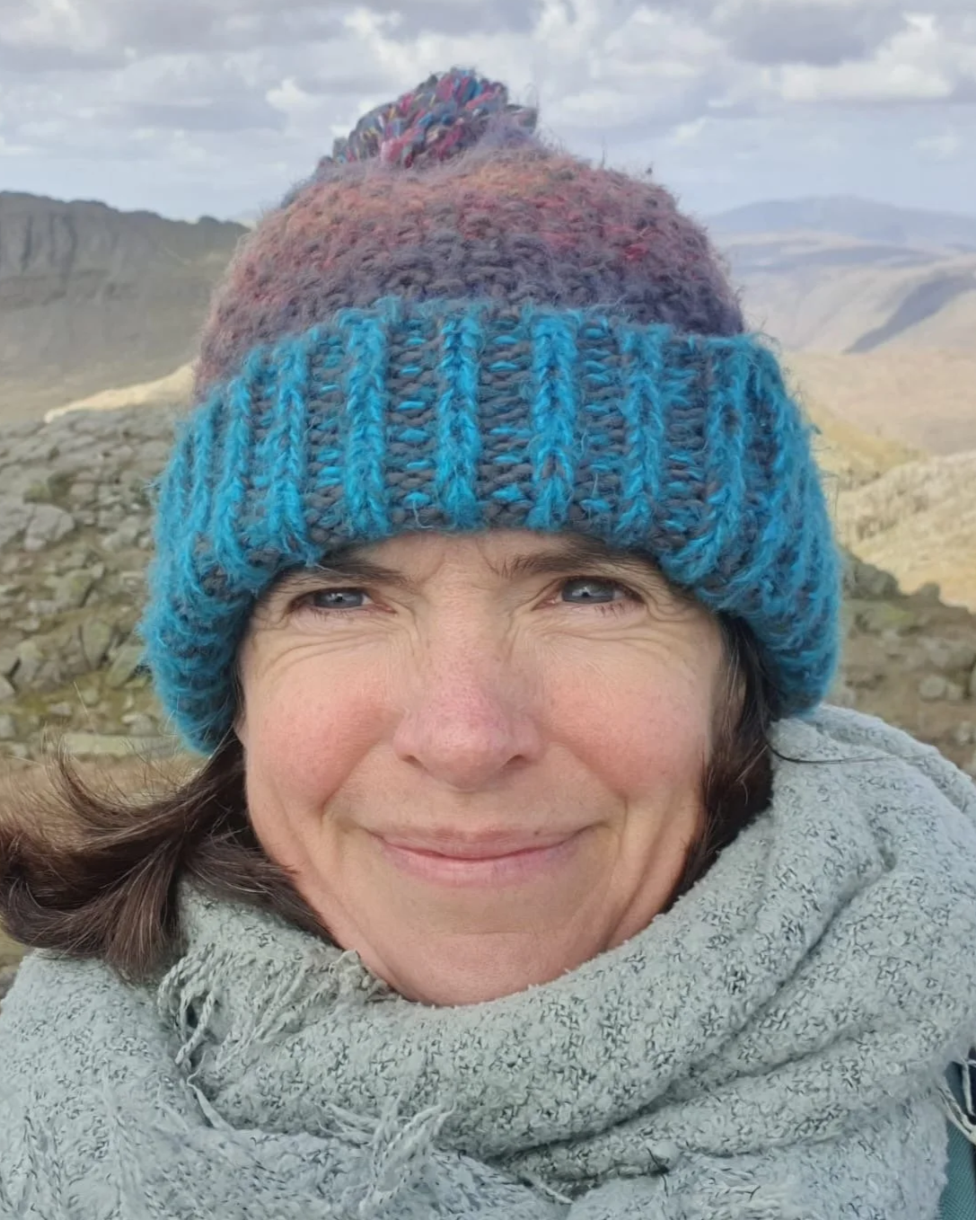 Close-up of a smiling woman wearing a colorful knit beanie and a gray scarf, outdoors in a mountainous landscape with cloudy sky.