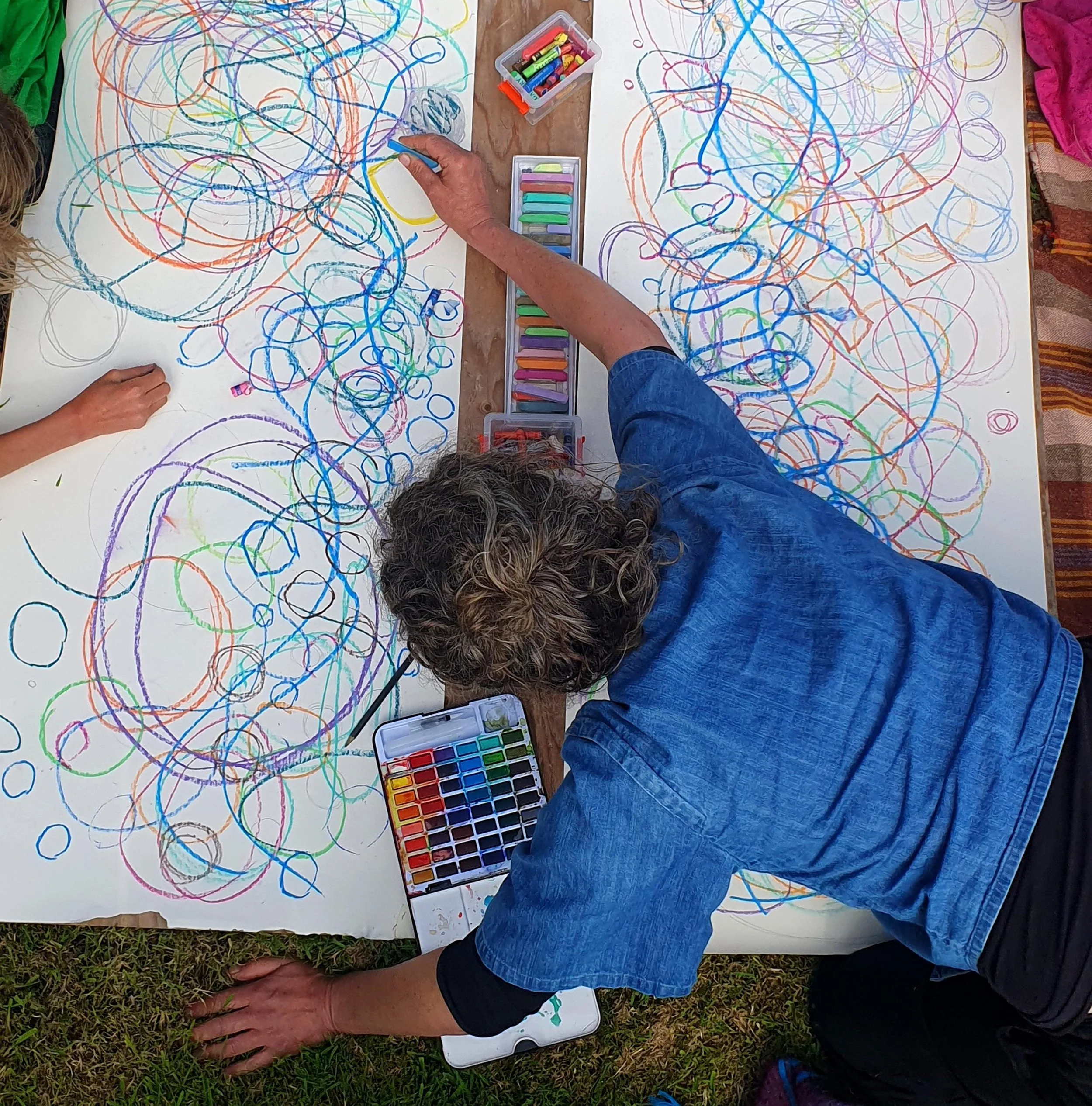 Children and adult drawing colorful abstract scribbles on large white sheets using pastels and markers, outdoors on grass.