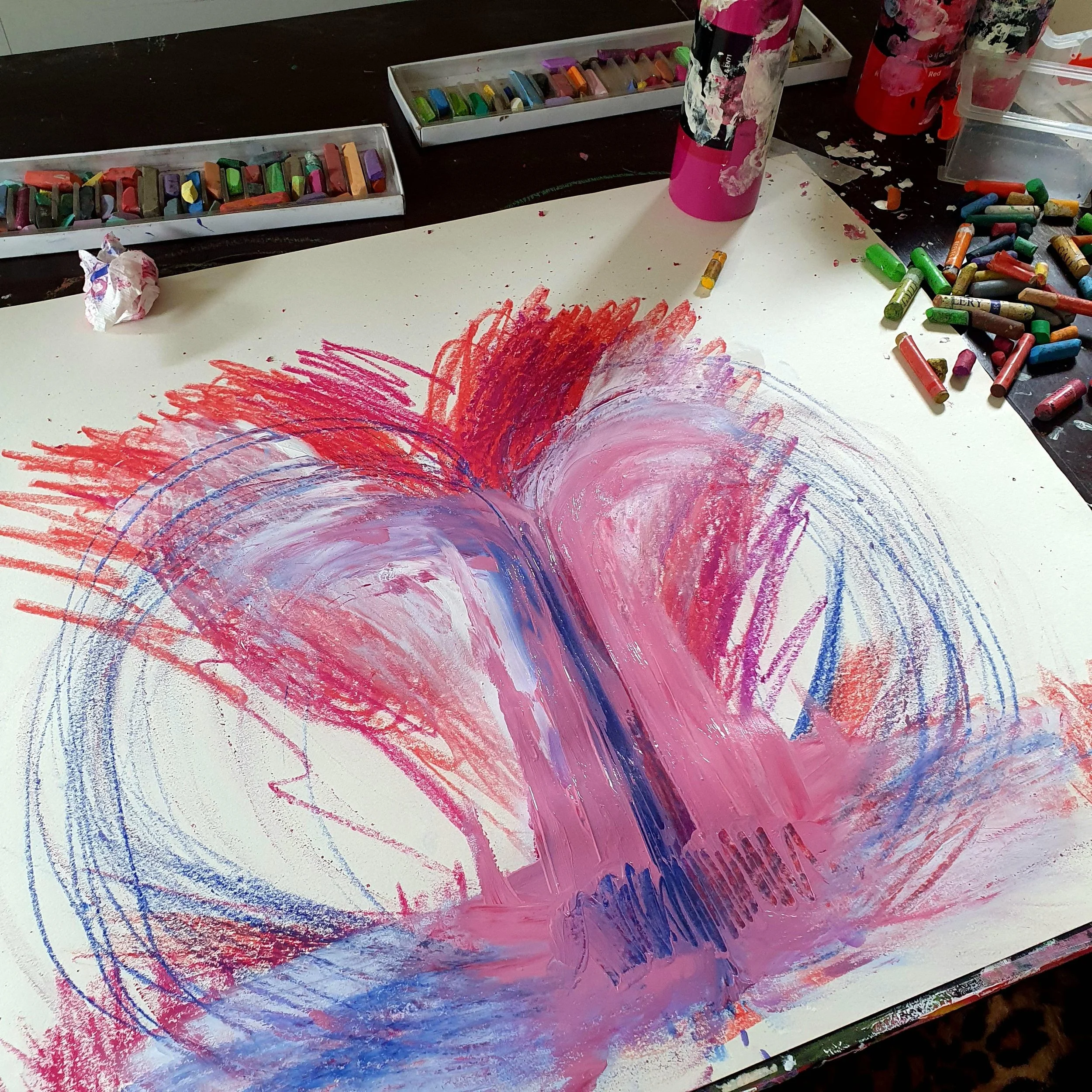 The results of an Embodied Drawing session showing the artwork with big red, pink, purple, and blue strokes on paper, surrounded by crayon boxes and containers on a dark wooden table.