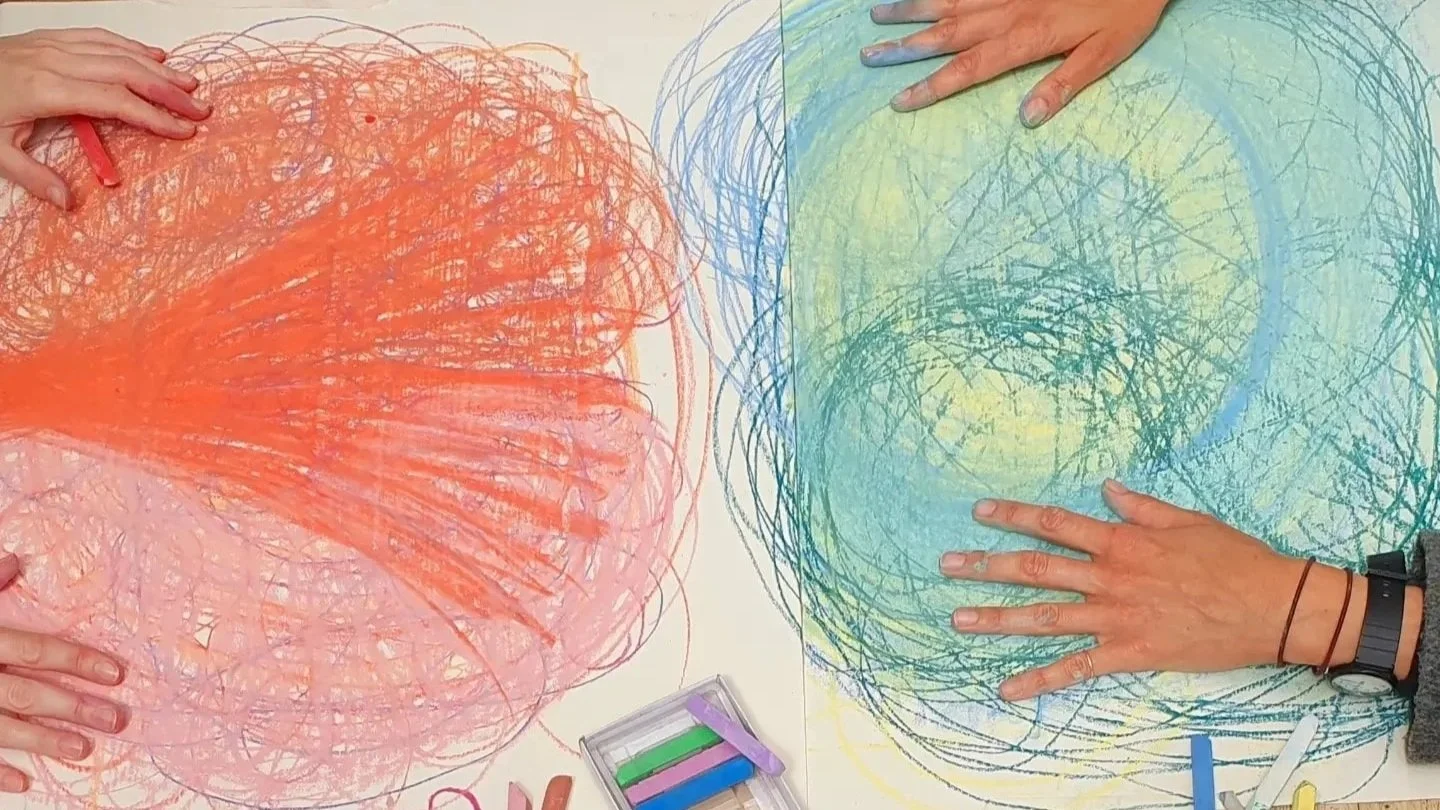 Two hands are pressed on large sheets of paper with colorful oil-pastel scribbles from an Embodied Drawing workshop.