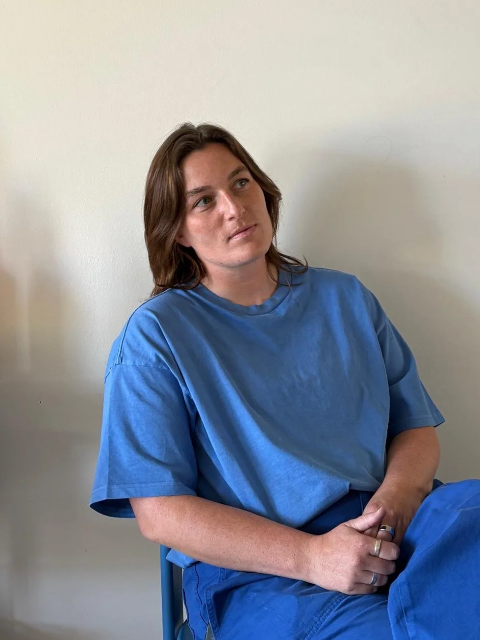 A woman with brown hair wearing a blue hospital gown sitting against a plain white wall, looking off to the side.