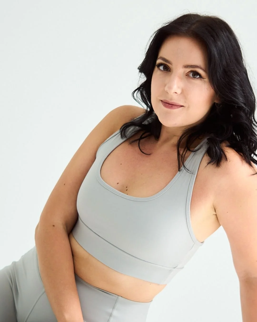 A woman with dark wavy hair wearing a gray sports bra and matching leggings, sitting against a plain white background.