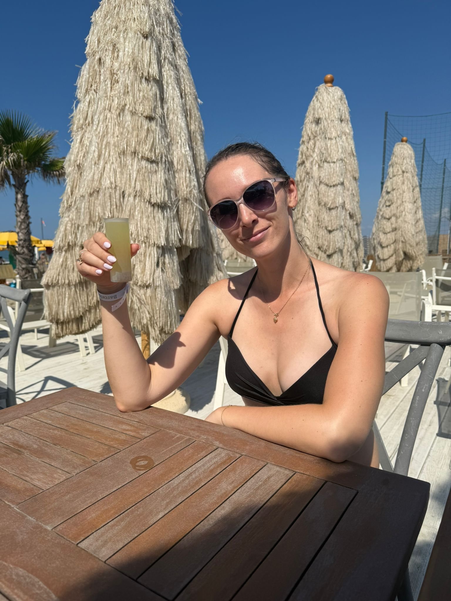 A woman with dark hair in sunglasses wearing a black bikini top sitting at an outdoor table at a sunny beach resort, holding a drink in her right hand, with straw umbrellas and a clear blue sky in the background.