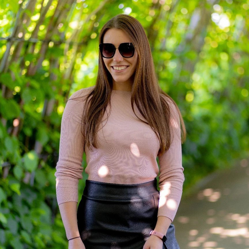 A woman with long brown hair, wearing large black sunglasses, a light pink long-sleeve shirt, and a black leather skirt, standing outdoors with green trees in the background.