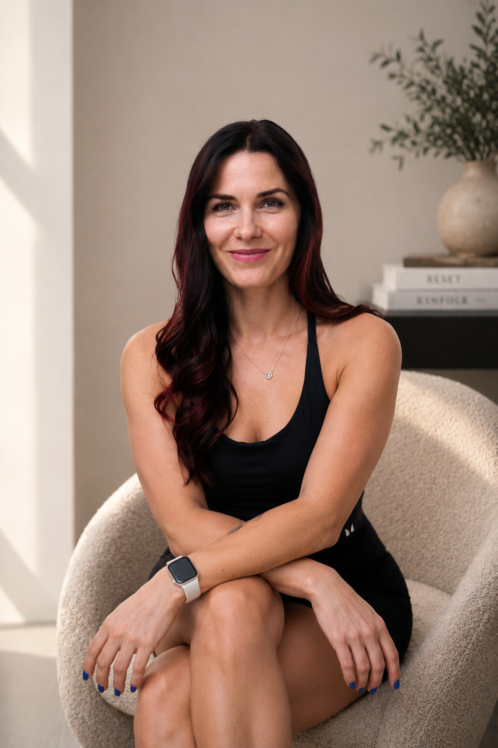 A woman with long dark hair and red highlights, sitting cross-legged on a beige armchair, smiling at the camera, wearing a black tank top, a smartwatch, and a necklace, in a cozy room with books and a plant in the background.