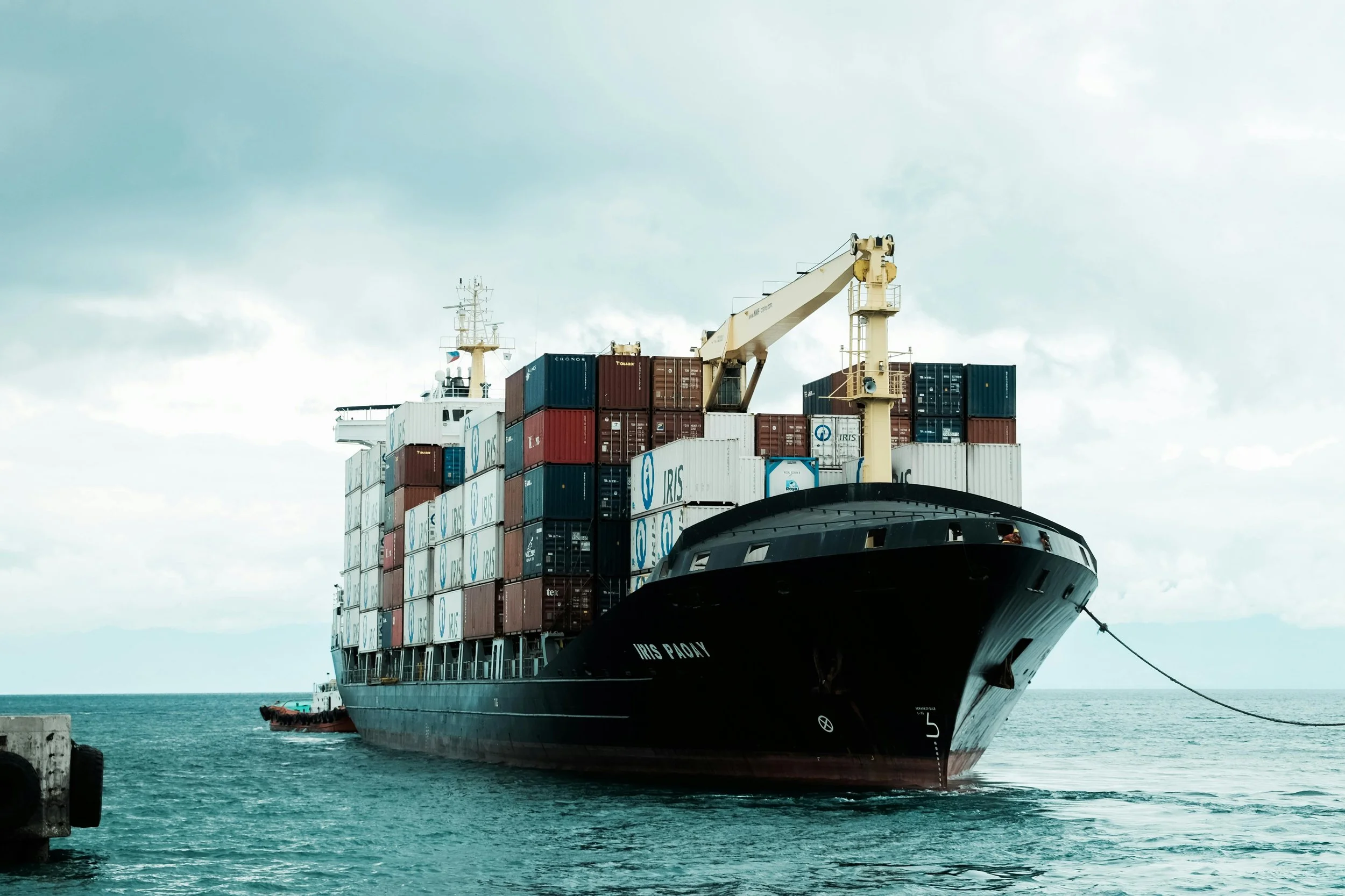 A large cargo ship transporting stacked shipping containers is docked at a port, with a crane on board for loading or unloading. The ship's name is Iris Pacy. The background shows cloudy skies at sea.