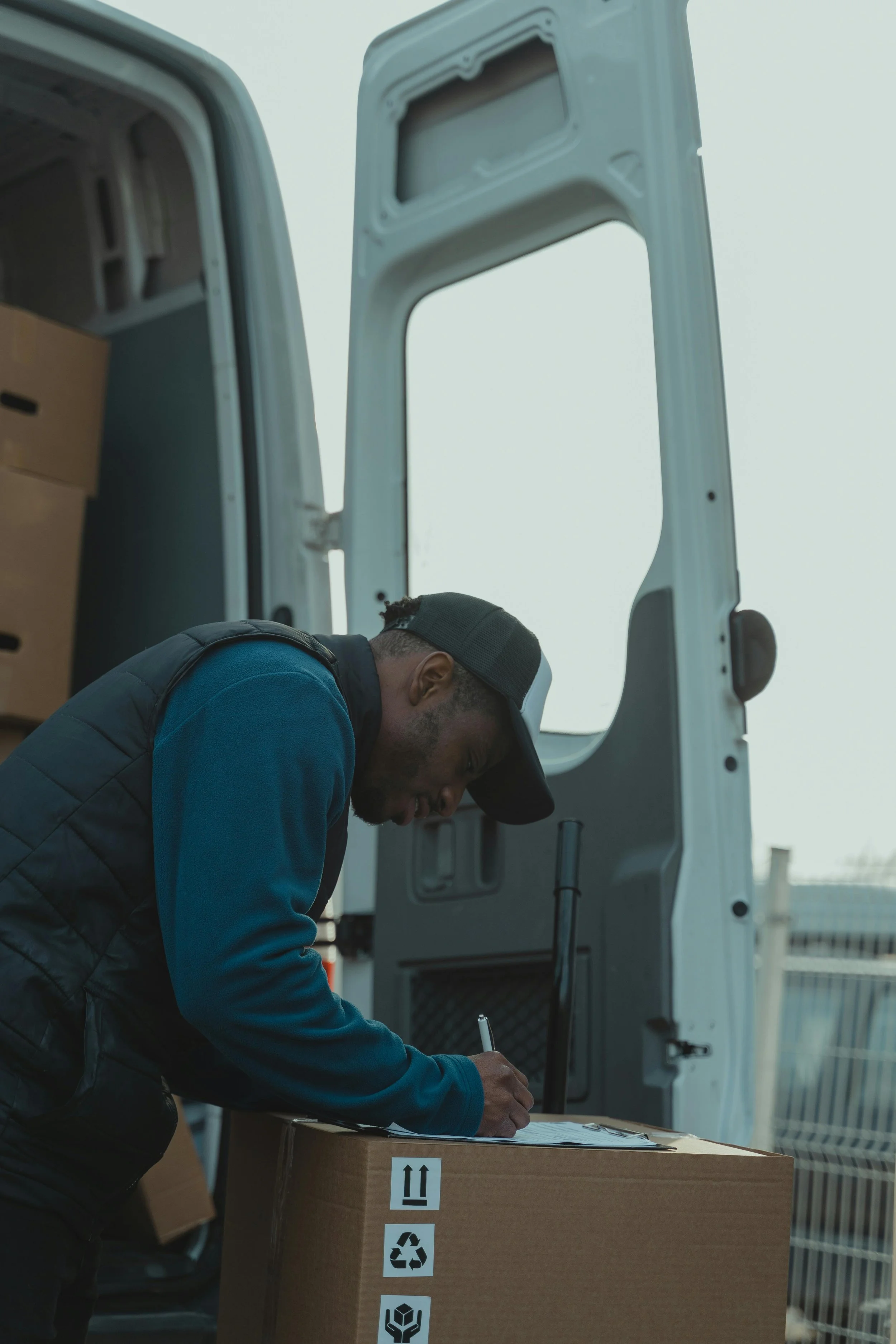 A man wearing a cap and vest is leaning over a large cardboard box with handling symbols, writing on a clipboard. He is beside an open van with a partially visible interior filled with boxes, parked outdoors.